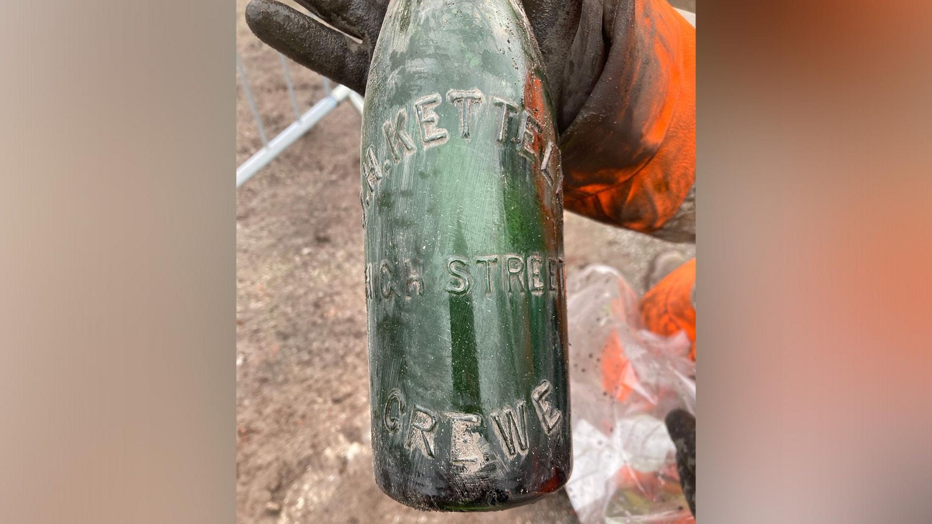 A green bottle is held up to the camera. It has the words High Street Crewe embossed on it