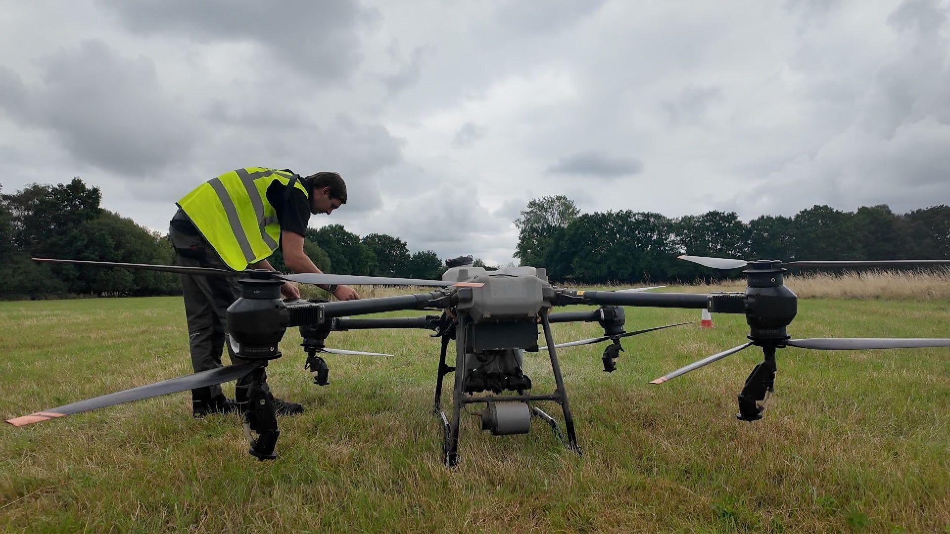 A man wearing a yellow fluorescent safety jacket leans over a large drone to fold out a rotor arm