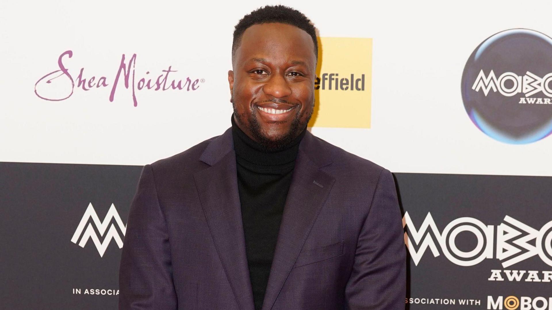 A man in a black suit jacket and black polo-neck stands in front of a black hoarding for the Mobo Awards. He has black cropped hair shaved around the sides. He is smiling.
