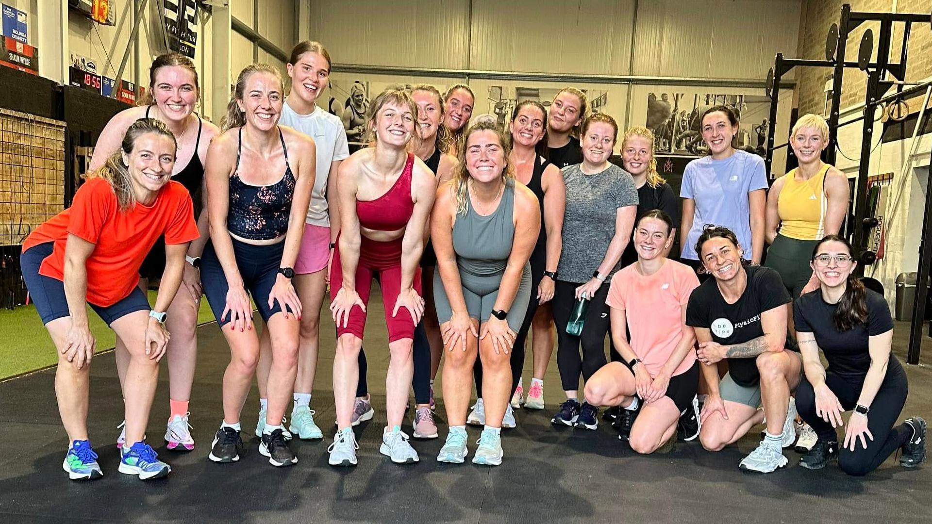 A group of 17 girls wearing active-wear pose for a picture in a gym after finishing a workout.