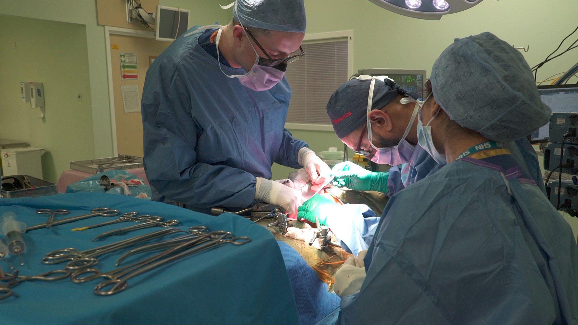 Three surgeons in blue scrubs at an operating table holding surgical instruments 