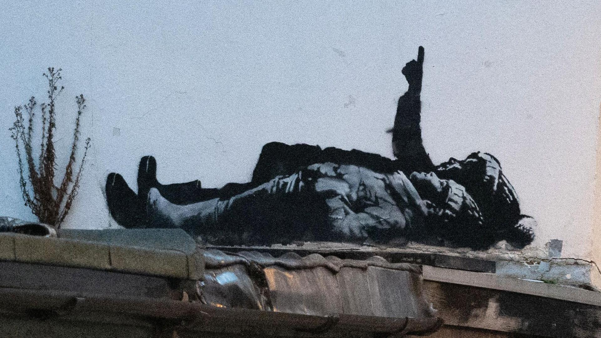 The black and white street art depicts two children lying on their backs and pointing upwards. The child nearest the camera is wearing a wooly hat with hands tucked into their jacket pockets. Behind them a larger person is pointing upwards in a bobbly hat (possibly a Santa type hat) also wearing boots.