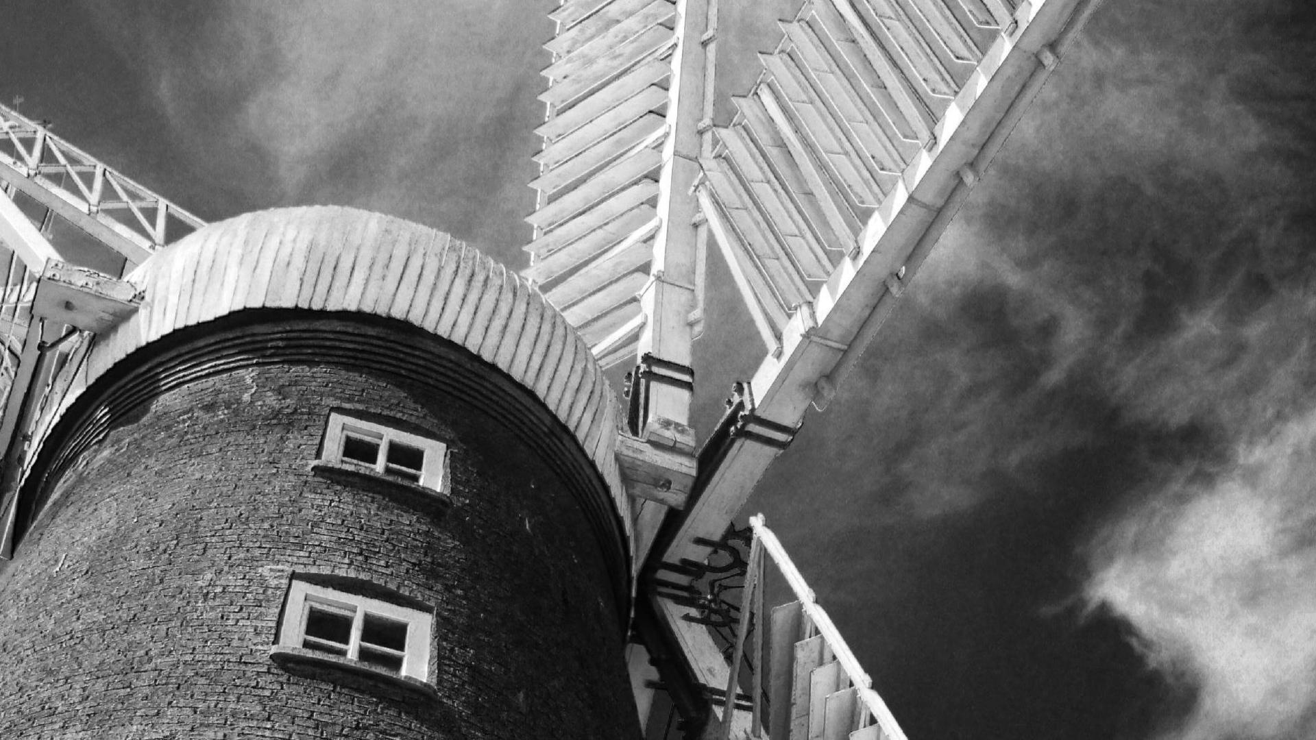 A black and white photo looking up towards the top of Alford Windmill, a tall, black building with small white windows, a white roof and large white sails.