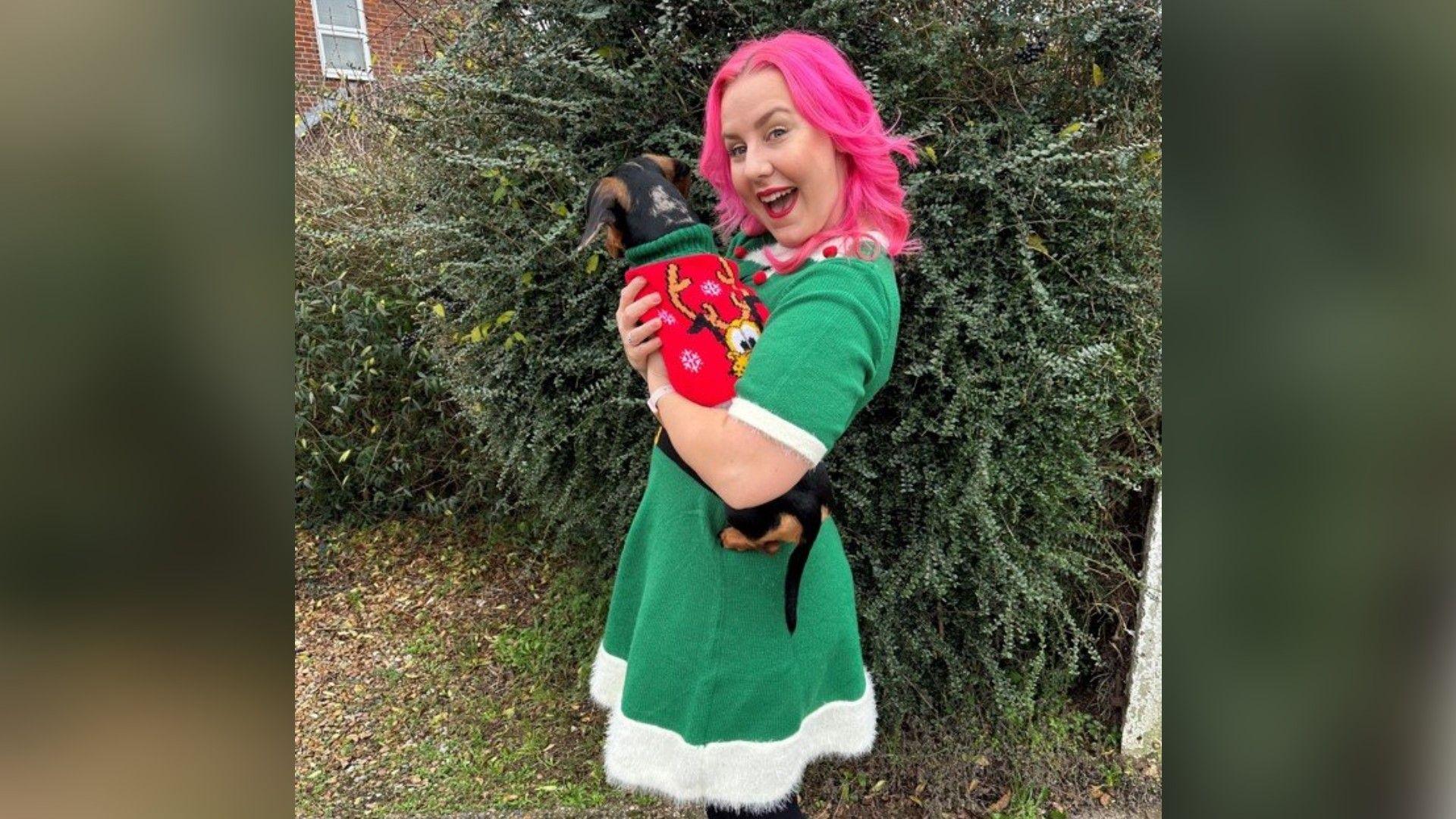 A smiling woman stood in front of a tree while holding a dog. She has pink hair and is wearing a green dress with a white trim. The dog is also wearing a red jumper with an image of a cartoon dog on.