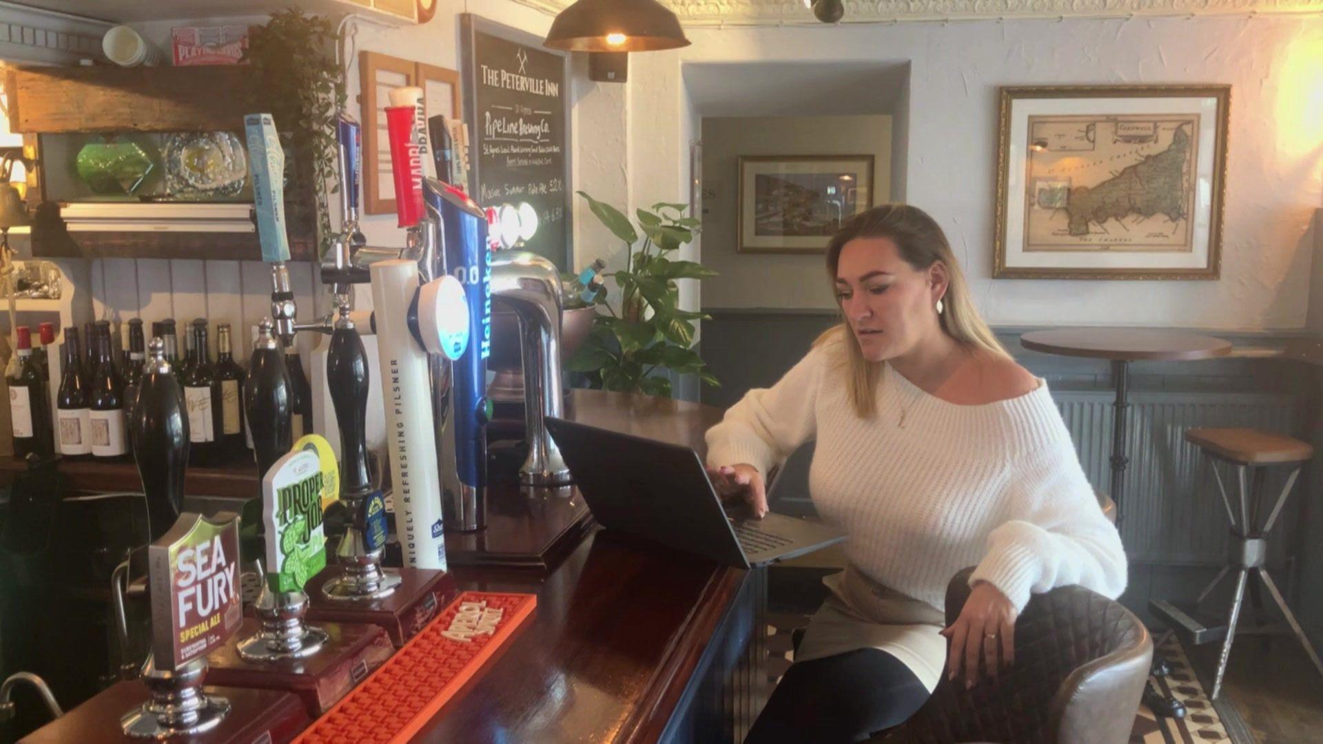 A woman with blonde hair, wearing a white jumper, on a laptop at a bar in a pub.