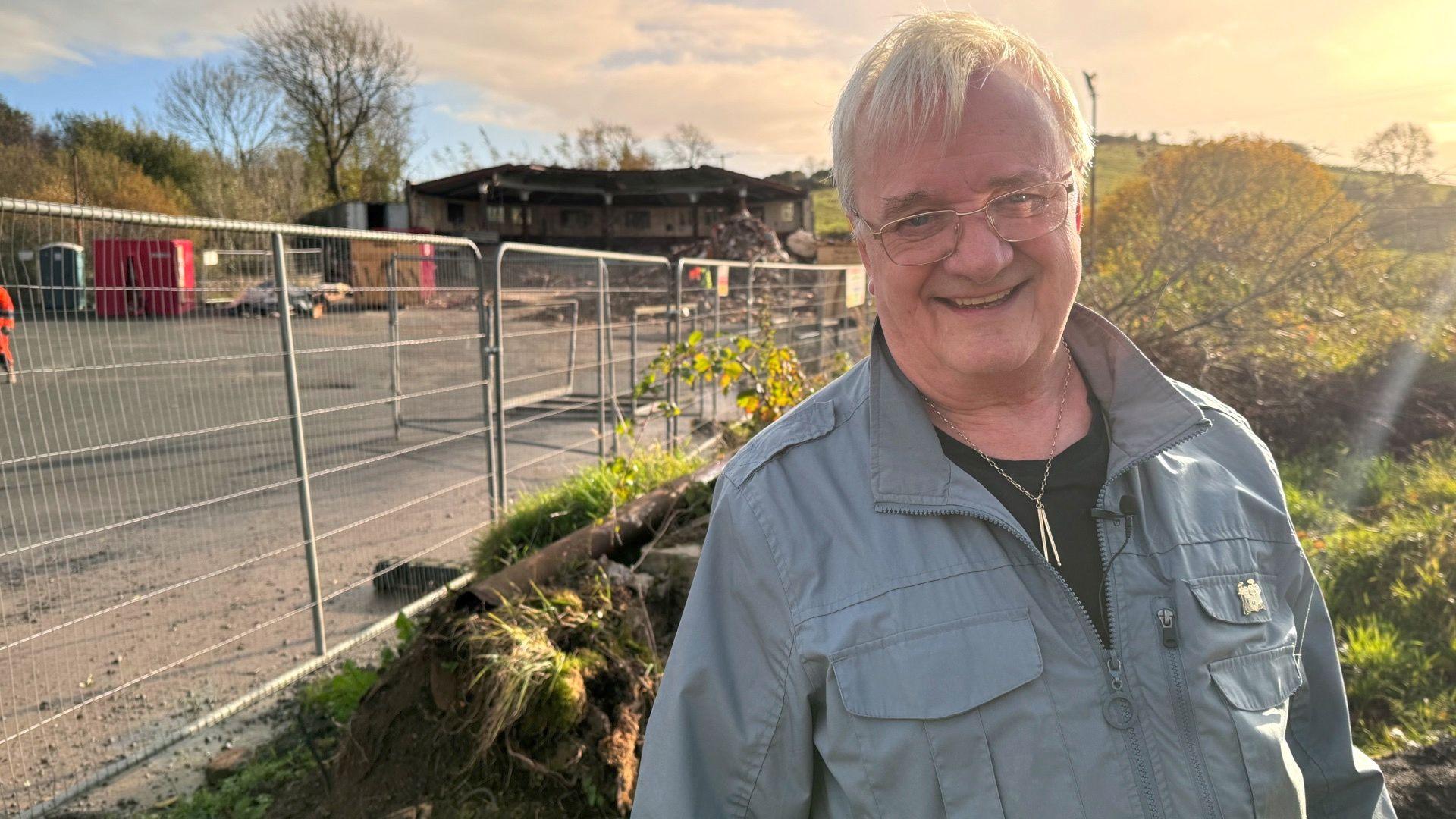 Vinny O'Donnell standing at the site, he's smiling at the camera with short white hair, he's wearing a black top, a light grey/blue jacket and a gold necklace with a gold pin on the jacket. He is wearing light framed glasses. Behind him are barriers and fields, the sun is shining from the right corner of the image. 