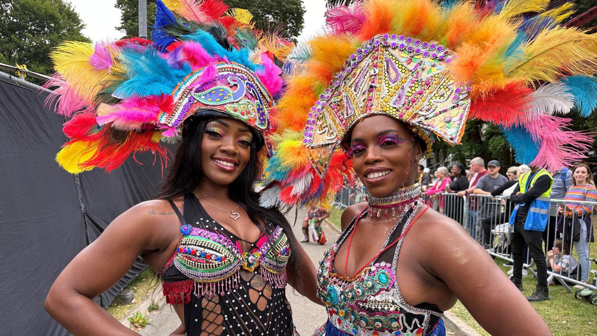 The smiling duo wears colourful headpieces and bodices adorned with feathers and gems. Their eye makeup is in green, blue and purple hues.