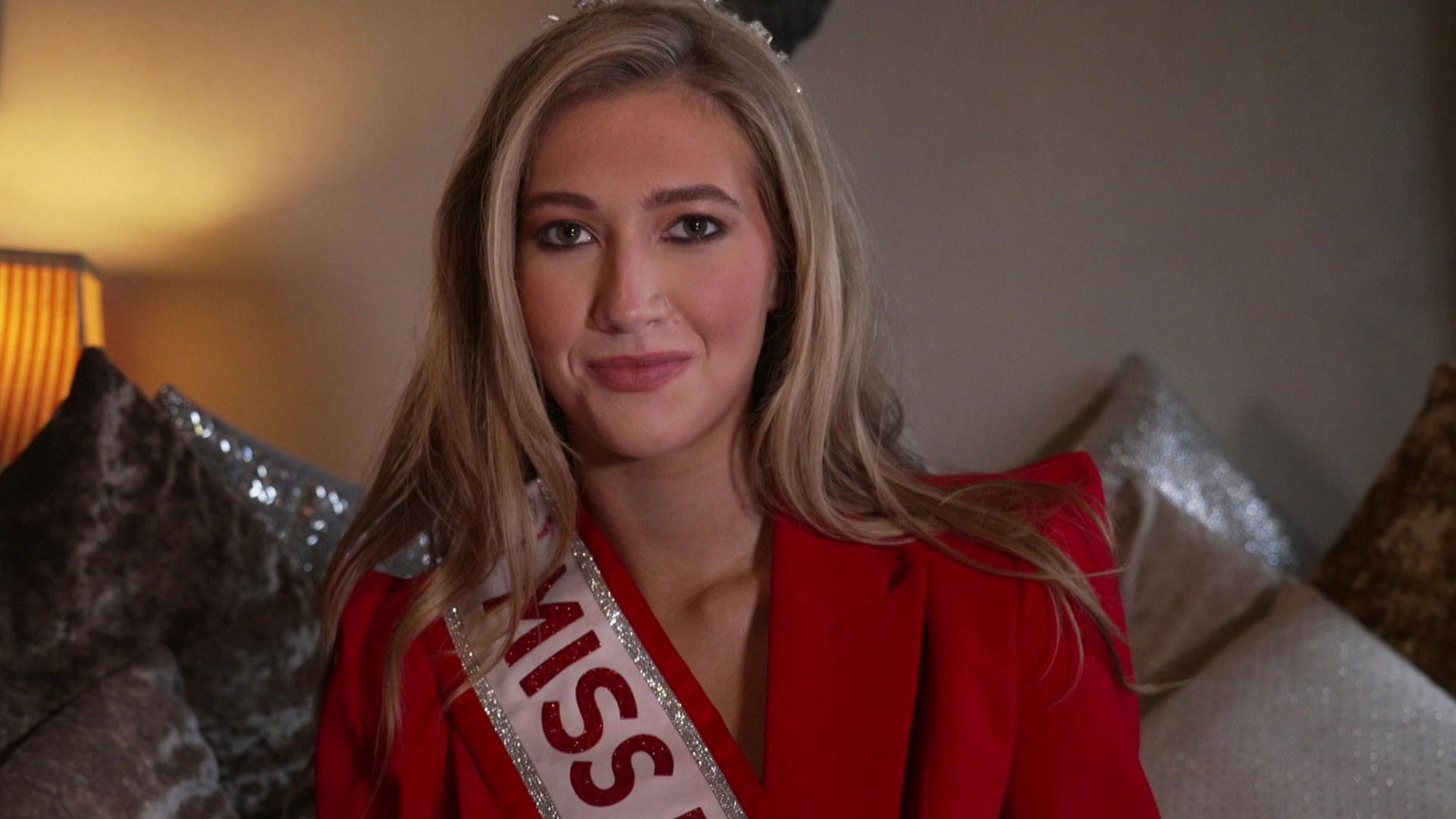 Tabitha sits on a couch surrounded by pillows. She has long blonde hair and wears a red suit, red and silver sash and silver crown.