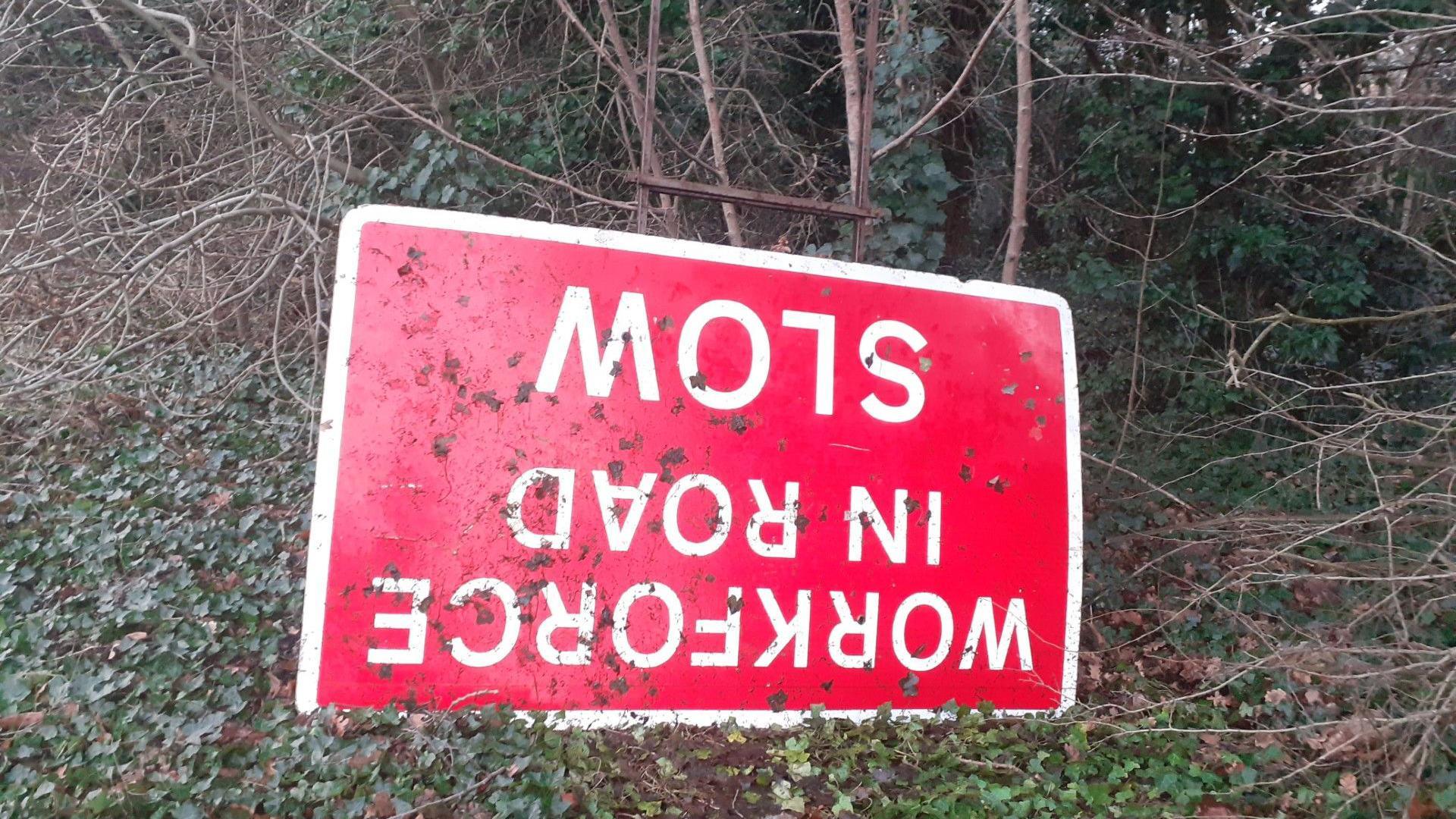 A discarded 'workforce in the road' sign, red background, rectangular shape with white writing, it was found discarded upside down on a verge by the roadside,