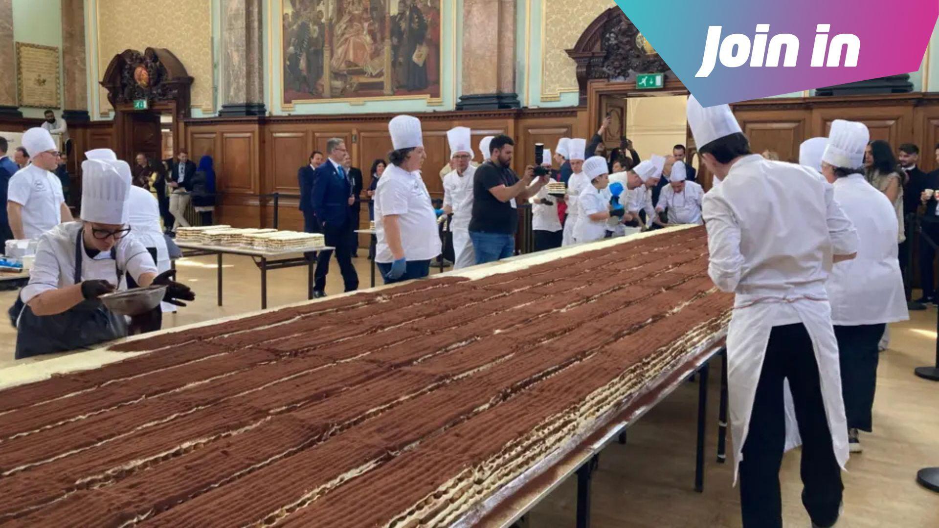 Chefs in white hats dust cocoa powder over a huge tiramisu spanning several tables.