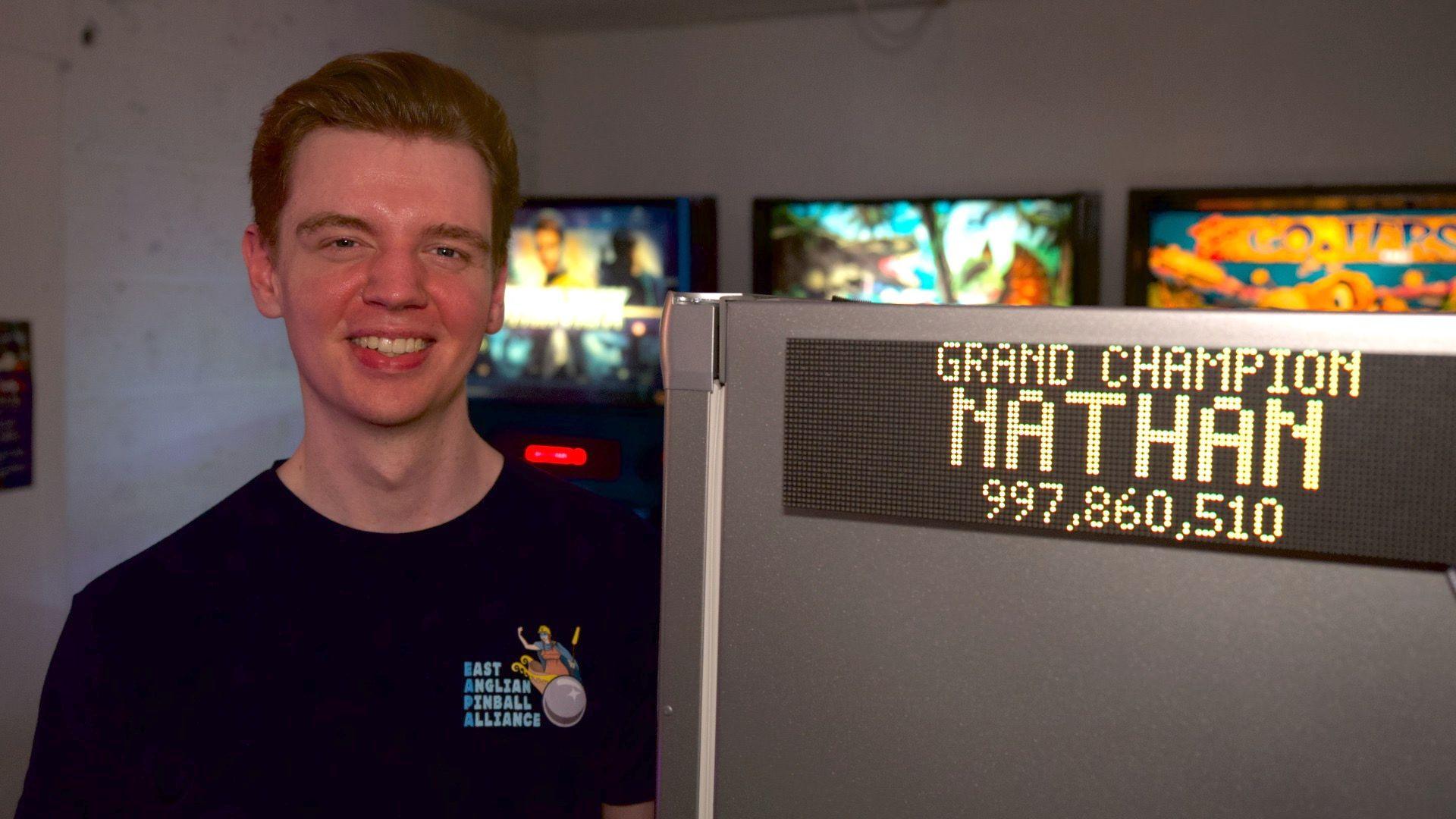 Nathan Garwood with short brown hair wearing an East Anglian Pinball Alliance tee-shirt. A sign next to him says "Grand Champion Nathan"