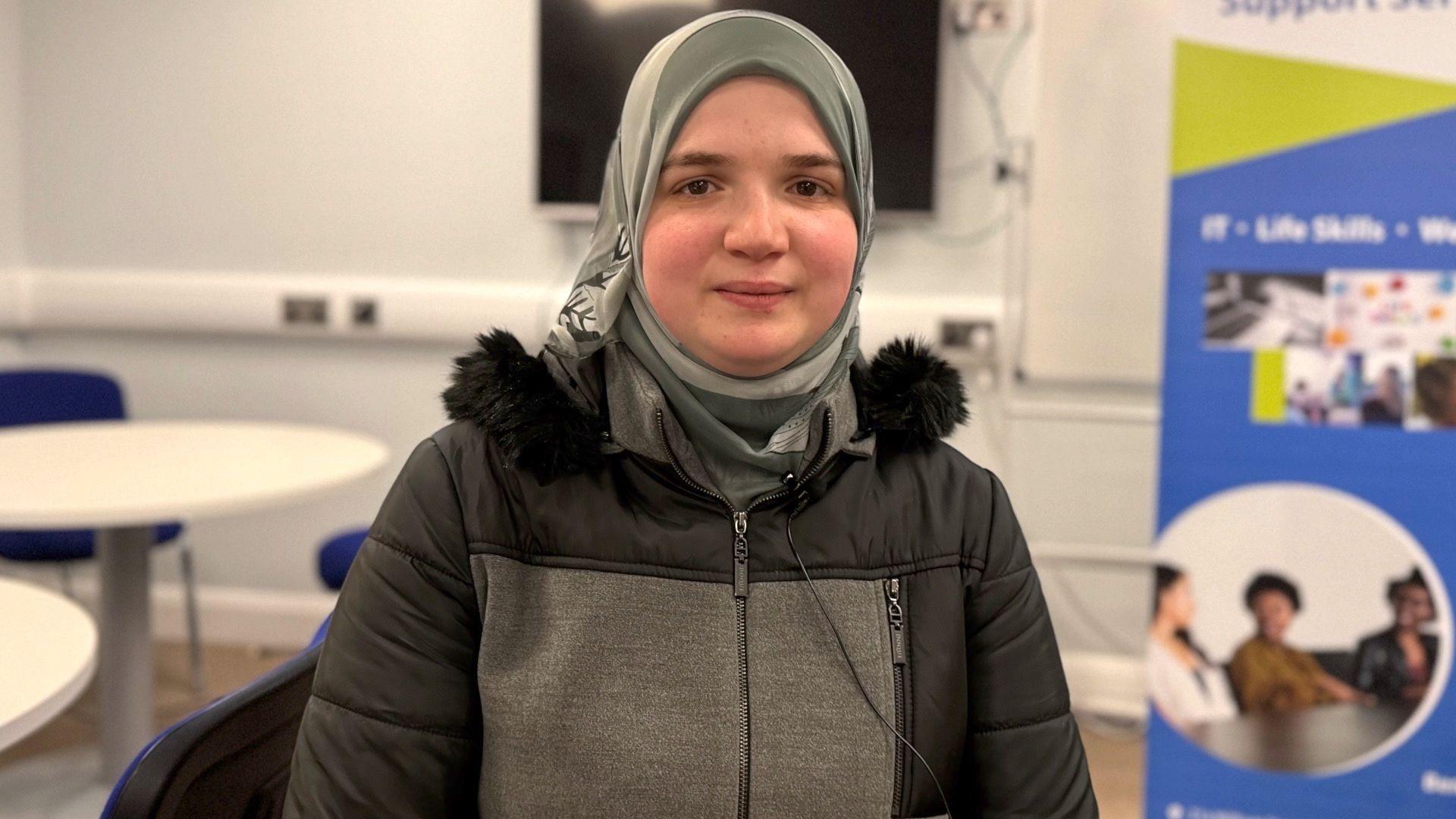 Sabah, she is wearing a grey headscarf and a black and grey coat and is sitting in a room with white walls. Behind her a white table and blue chair can be seen.