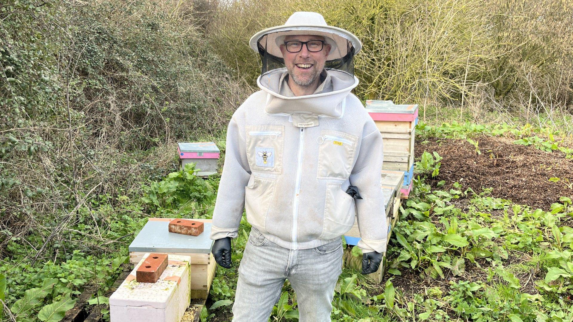 Burrë që fërkon një kapelë dhe xhaketë të mbuluar me rrjetë për mbajtjen e bletëve, duke qëndruar pranë një numri kosheresh bletësh.