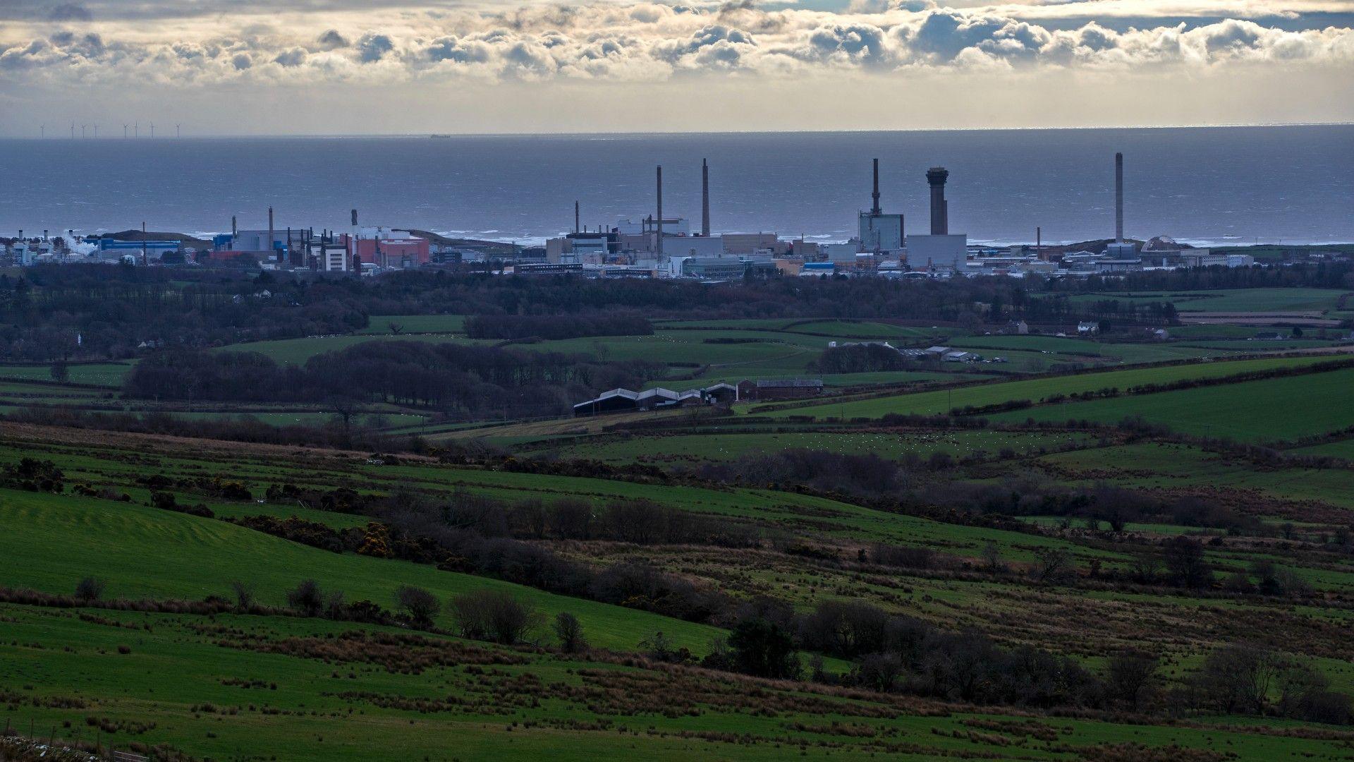 A general view of the Sellafield site seen from a distance, many chimneys are high over the site. There is the sea in the background and the clouds sit on the horizon.