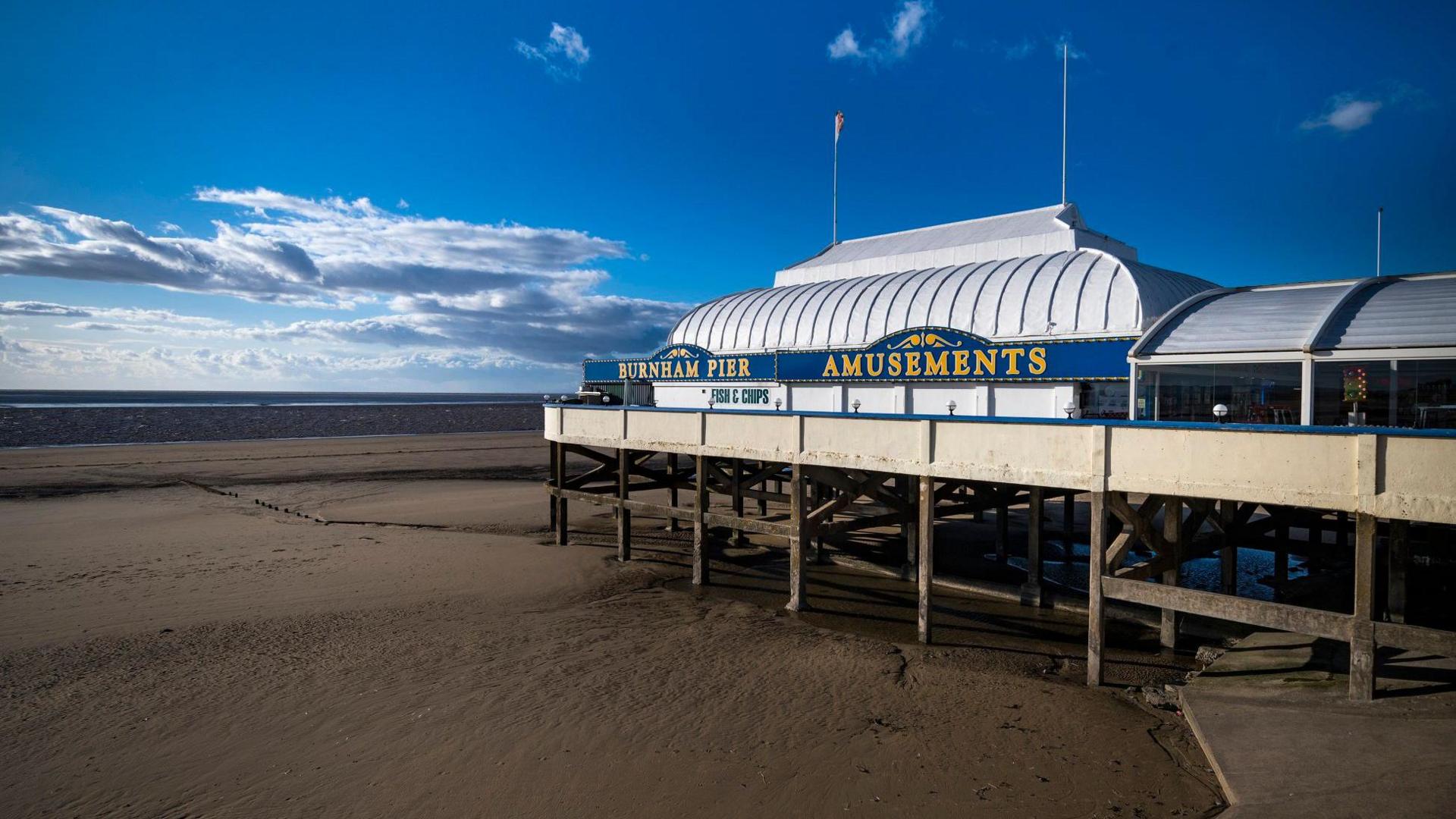 Burnham-on-Sea pier is seen at low tide, with mud flats underneath it. On the side of the structure is written "Burnham Pier Amusements" in yellow letters on a blue background. The skies overhead are clear and blue