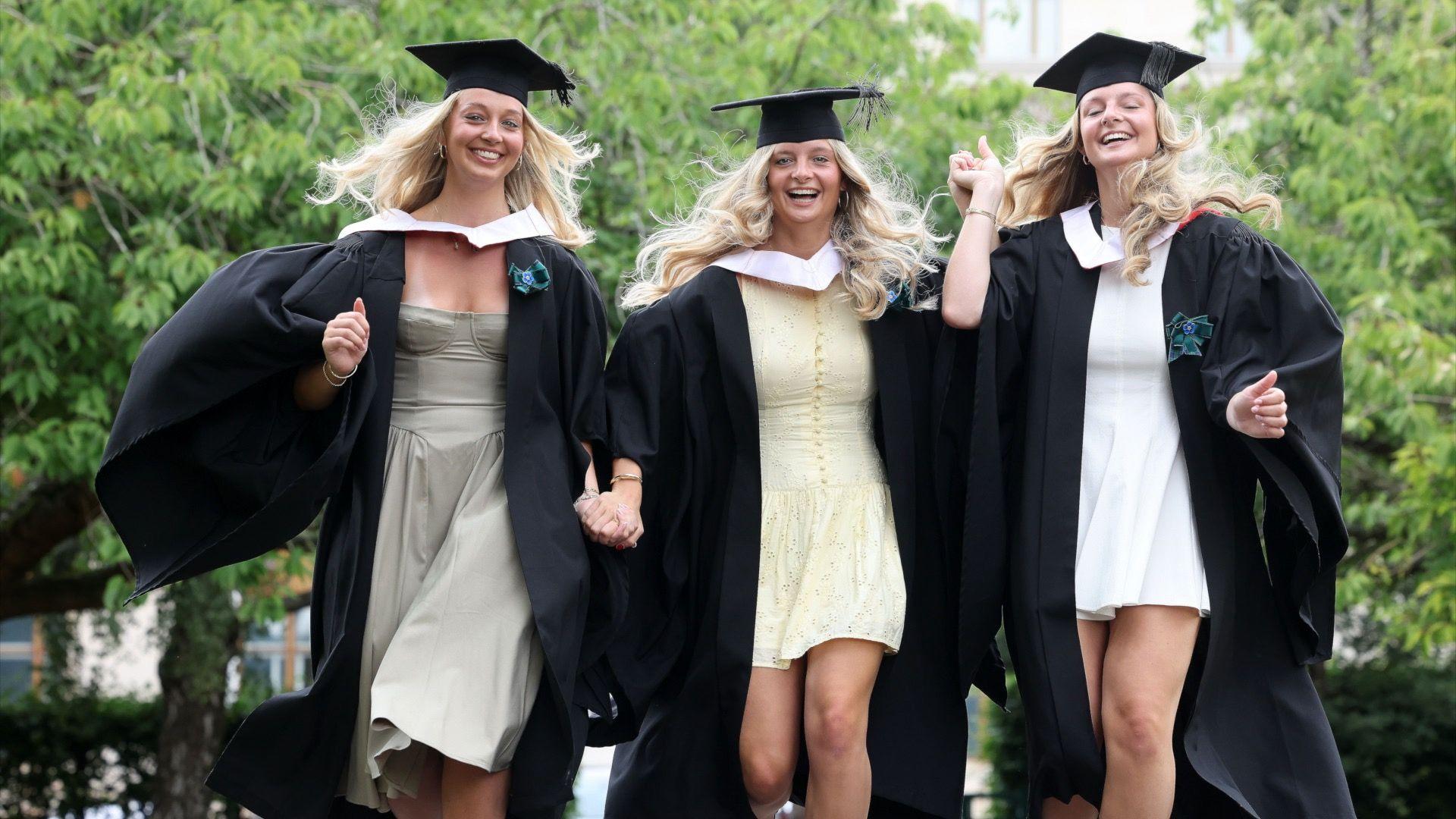 Three young woman with blonde hair in cream dresses and graduation gowns and caps walk towards the camera smiling