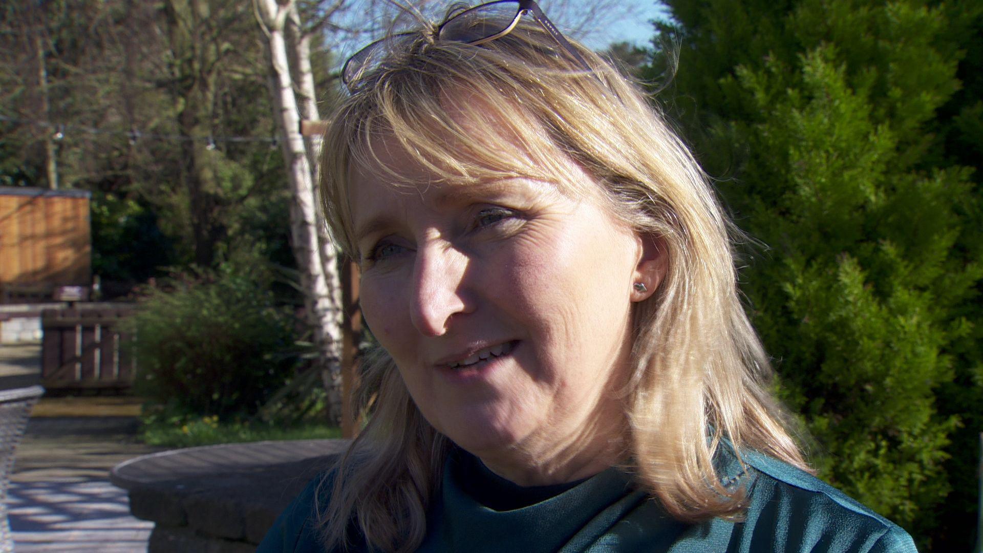 Sandra Peake. She has shoulder-length blonde hair with a fringe, wearing glasses on top of her head, a blue top, and stud earrings.