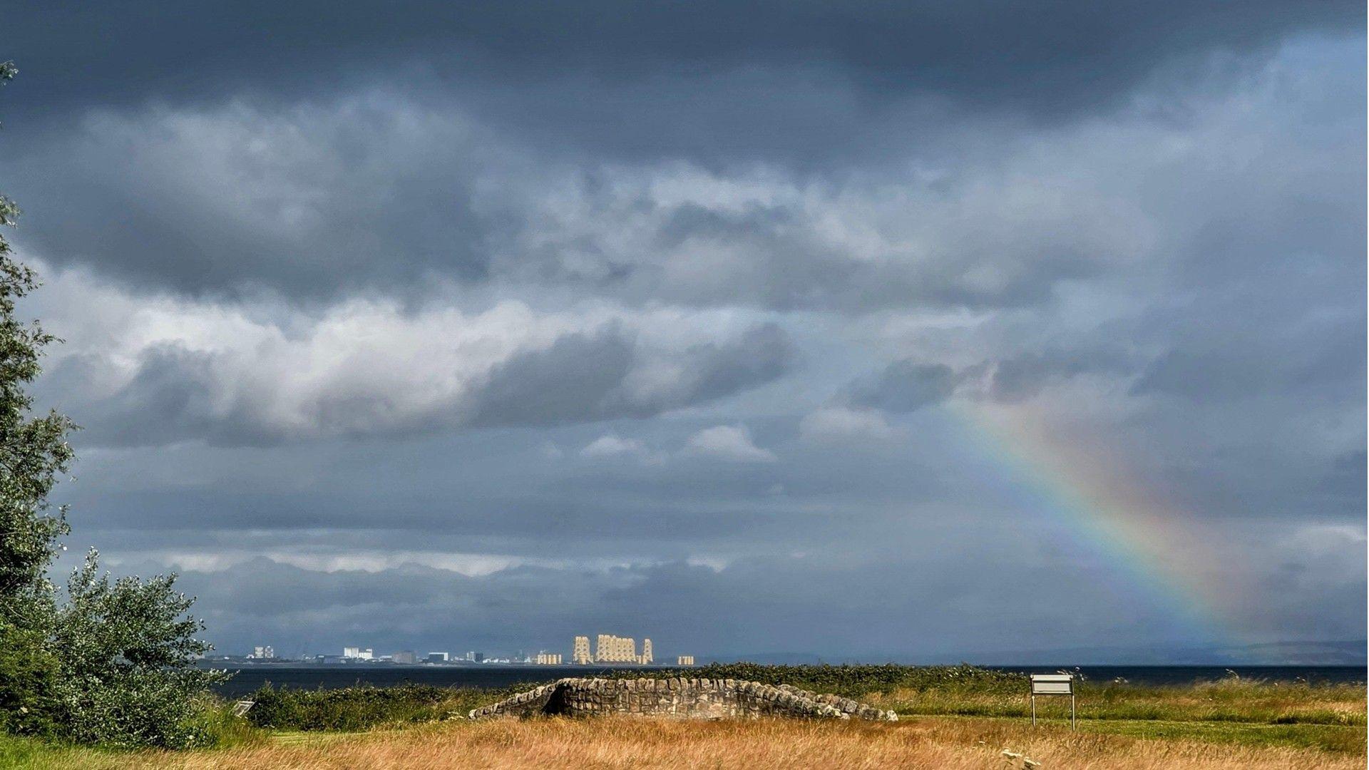 Plenty of cloud and a rainbow over East Lothian, Scotland. There is yellow grass in the foreground and tall cream-coloured buildings in the distance. 