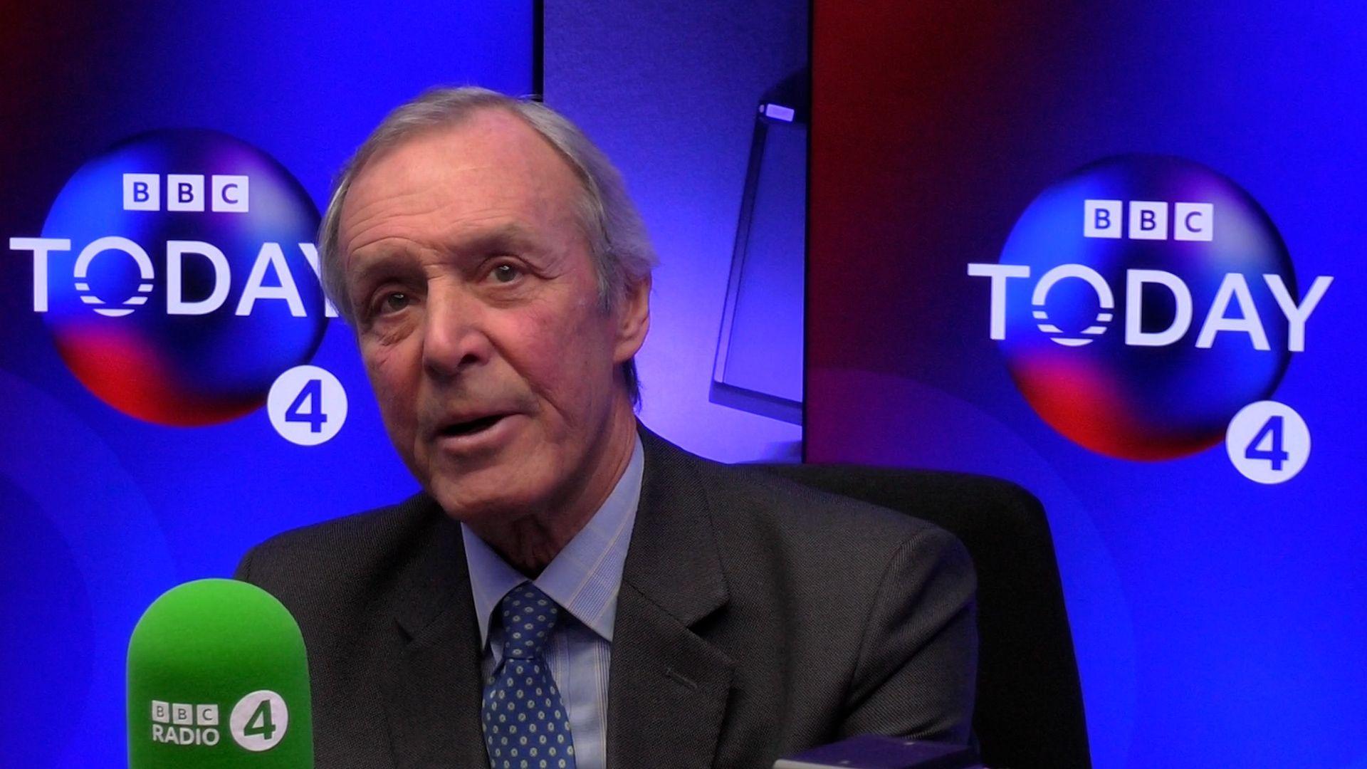 A man in a suit with short white hair is sat in a radio studio. He is speaking into a microphone labelled "BBC Radio 4"