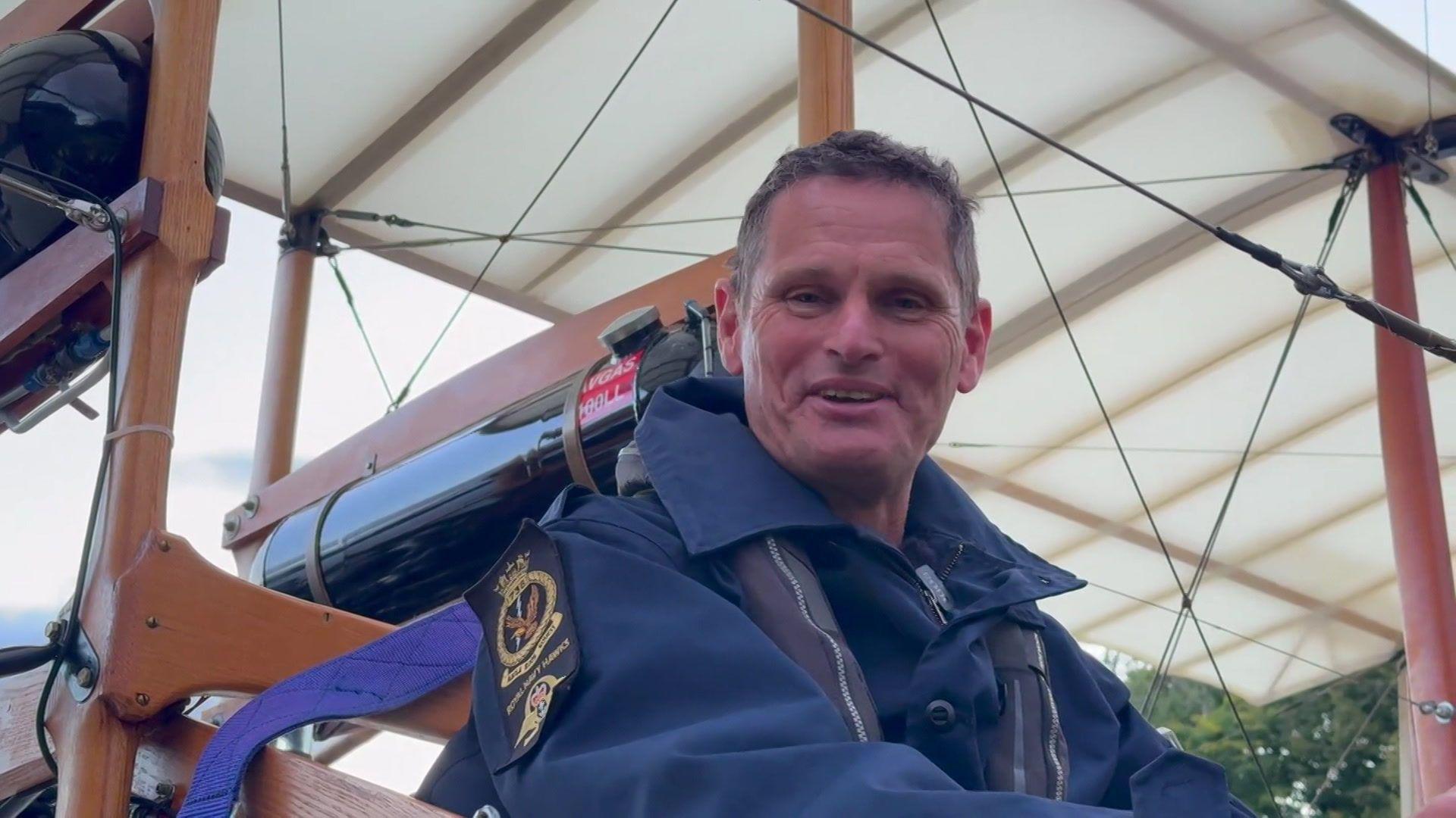 Lieutenant Commander Götke at the cockpit of Waterbird. The image is a close-up of him speaking to the camera. He is wearing blue overalls and the structure of the aircraft can be seen behind him.