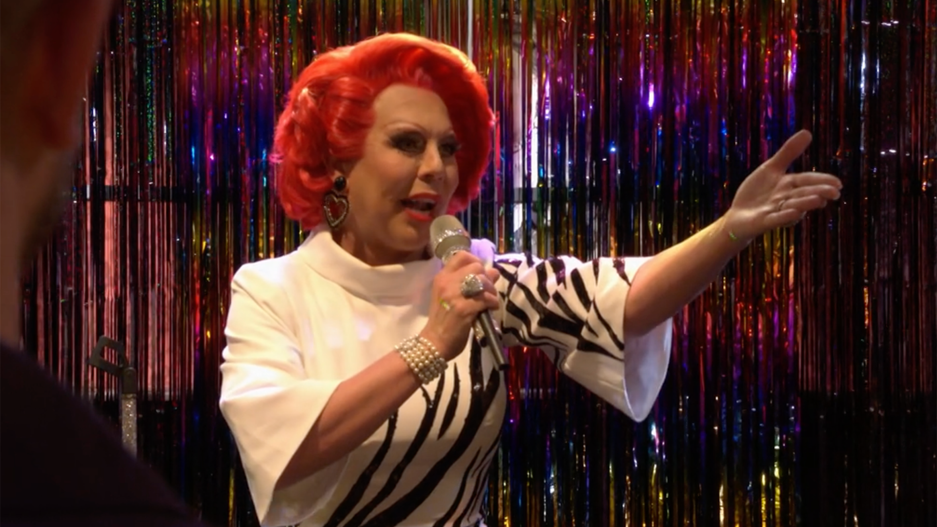la voix, a drag performer in a red wig and white tiger print top with wide sleeves, is holding a microphone in front of a glittery curtain. with her left hand she gestures to the audiences