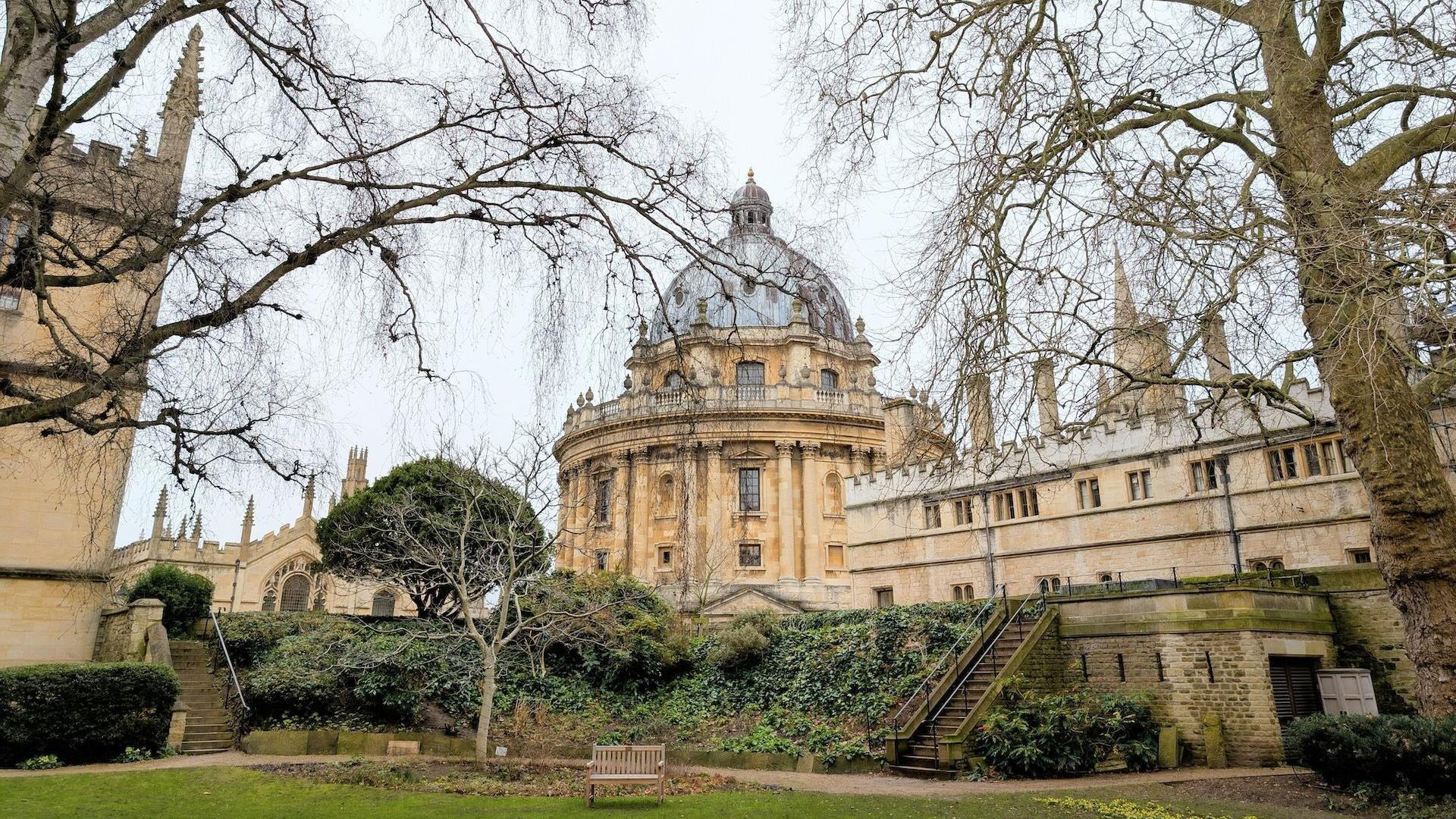 Stone buildings behind a park. The central building has a doomed roof. The stone is a pale yellow. In the foreground there is an area of grass and leafless trees. 