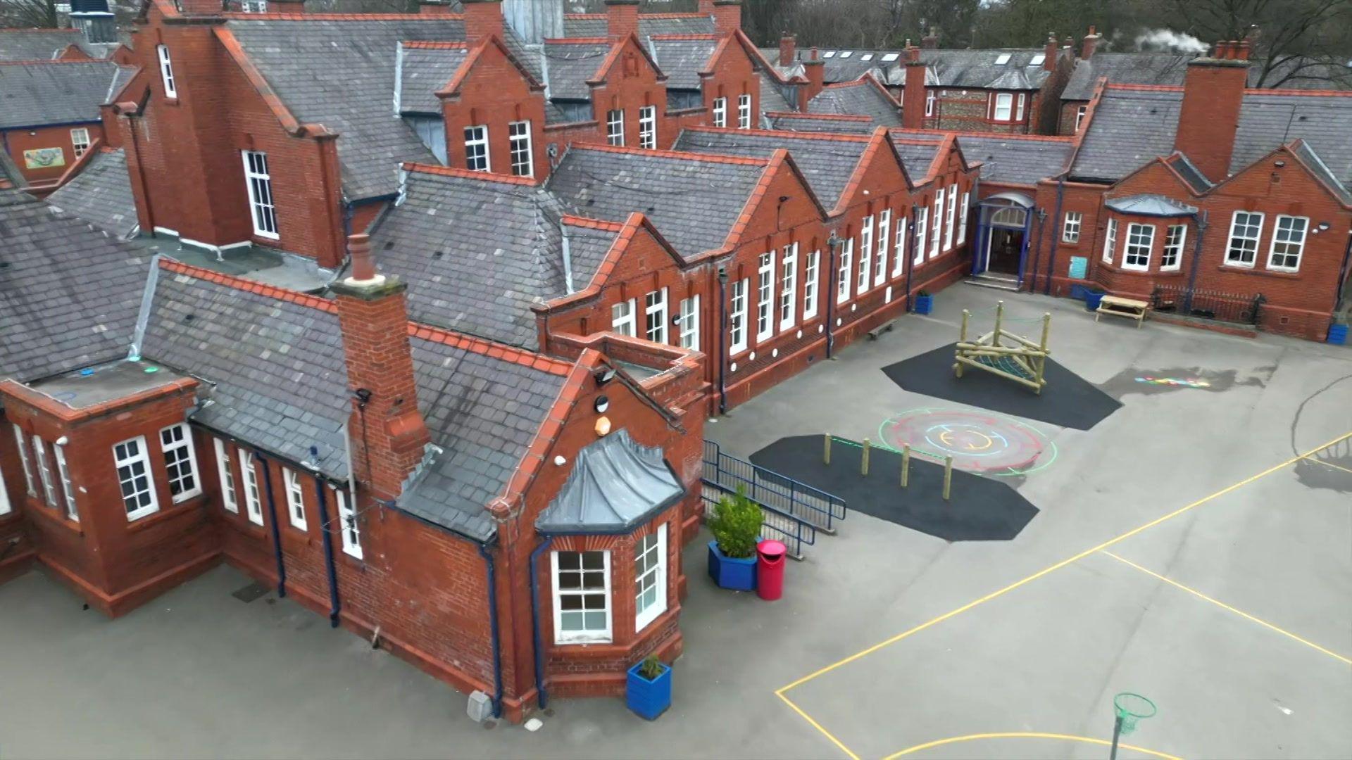 A large red brick building with chimney turrets and white bay windows with a school playground in front of it