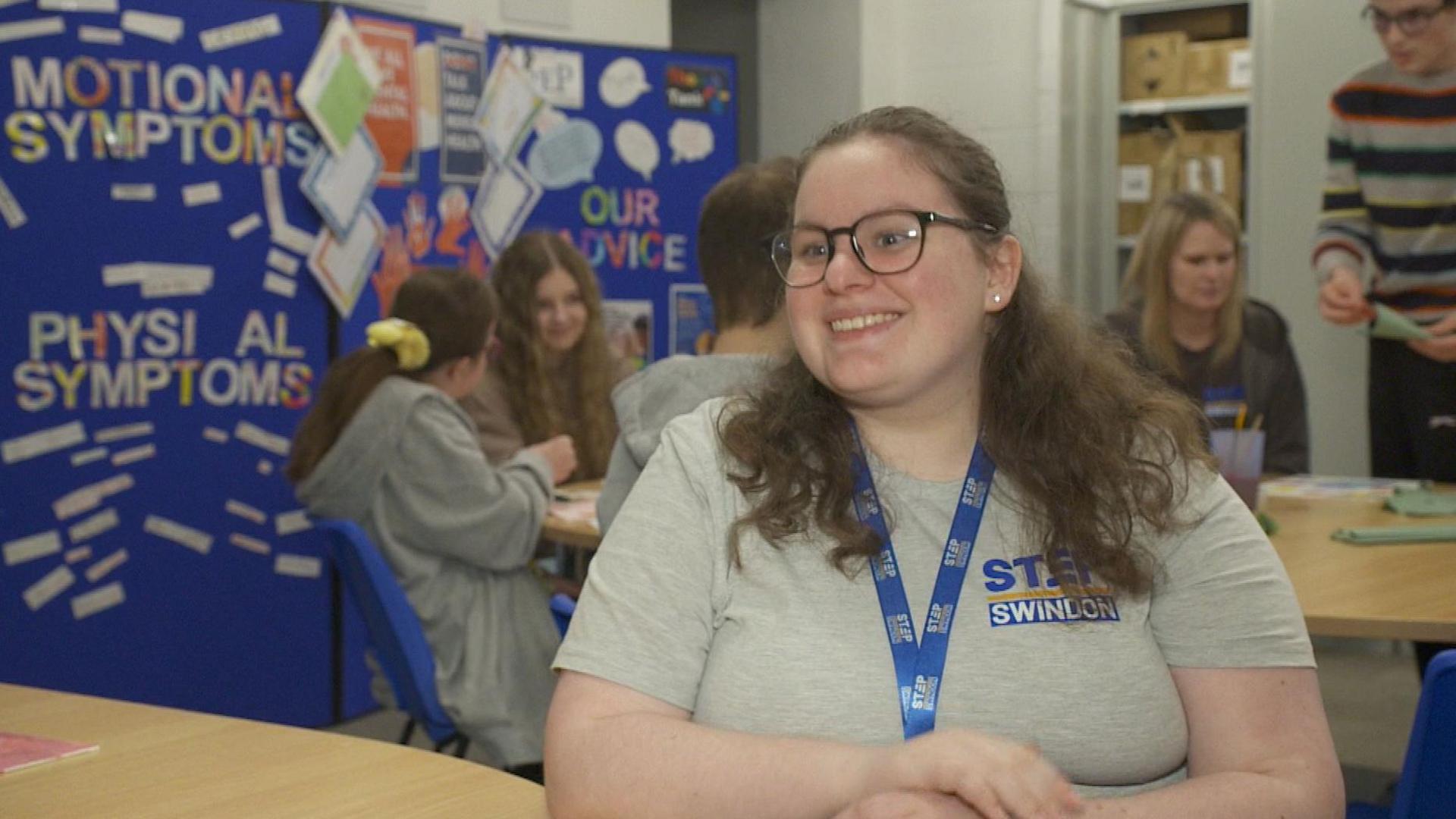 Jaime wears glasses and has long curly hair. She is smiling for the camera and wearing a lanyard and t-shirt with the STEP Swindon logo on. She is in a classroom-like environment with young people behind her.