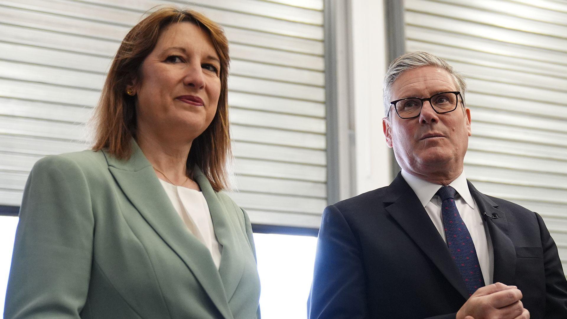 An image of Rachel Reeves on the left in a grey suit, and Keir Starmer on the right in a black suit, stood in front of windows with closed blinds, during a visit to Horiba Mira in Nuneaton in June 2025.