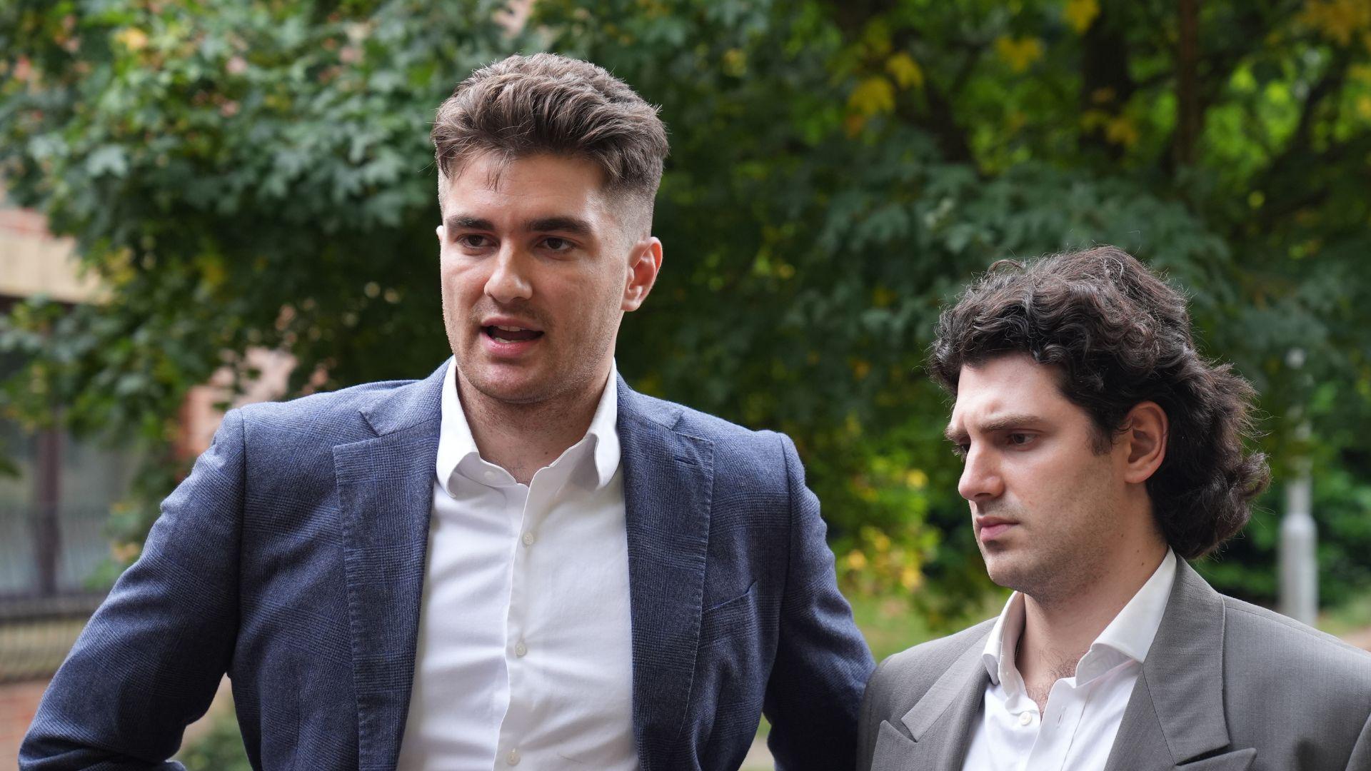 Two young men stood outside in front of some trees. On the left, a man wearing a blue suit jacket and white shirt. he has short brown hair. On the right, a man in a grey suit jacket and white shirt with long curly brown hair