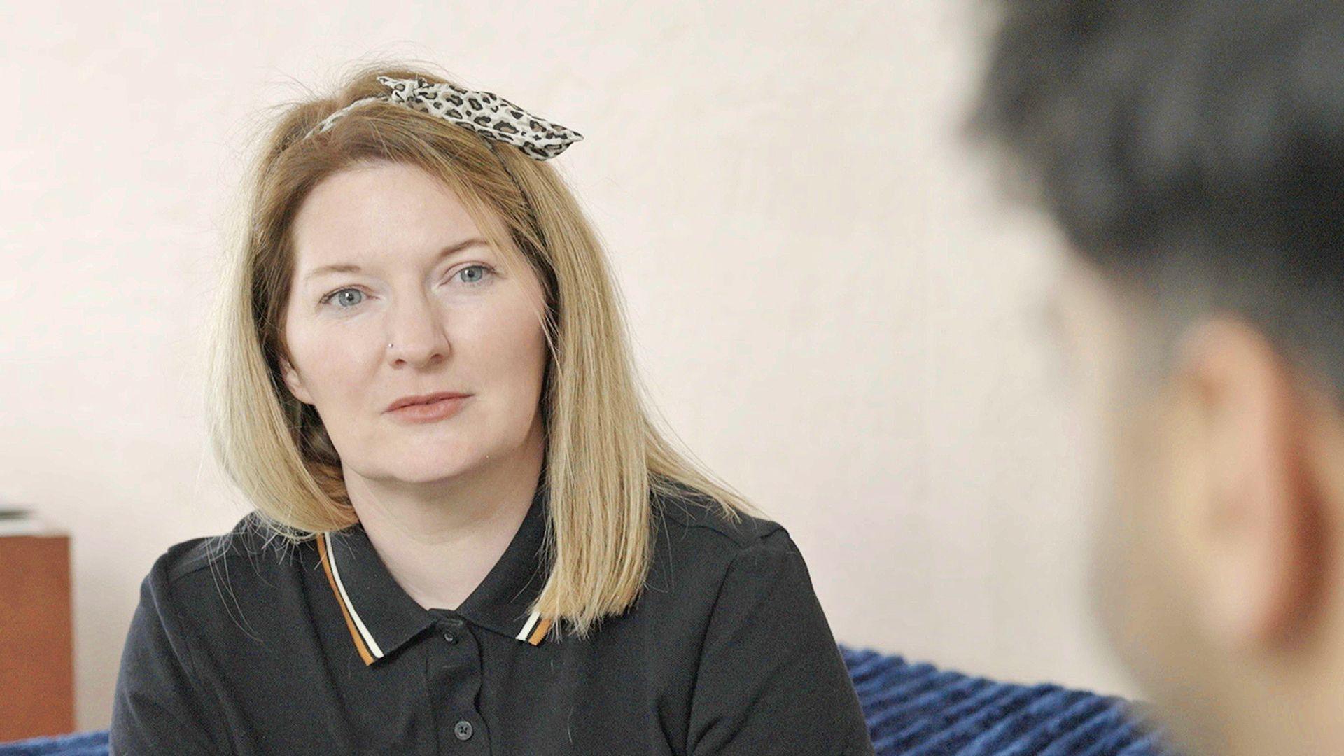 A woman looking towards the camera, wearing a black polo t-shirt, with straight fair hair and a leopard-print head band.