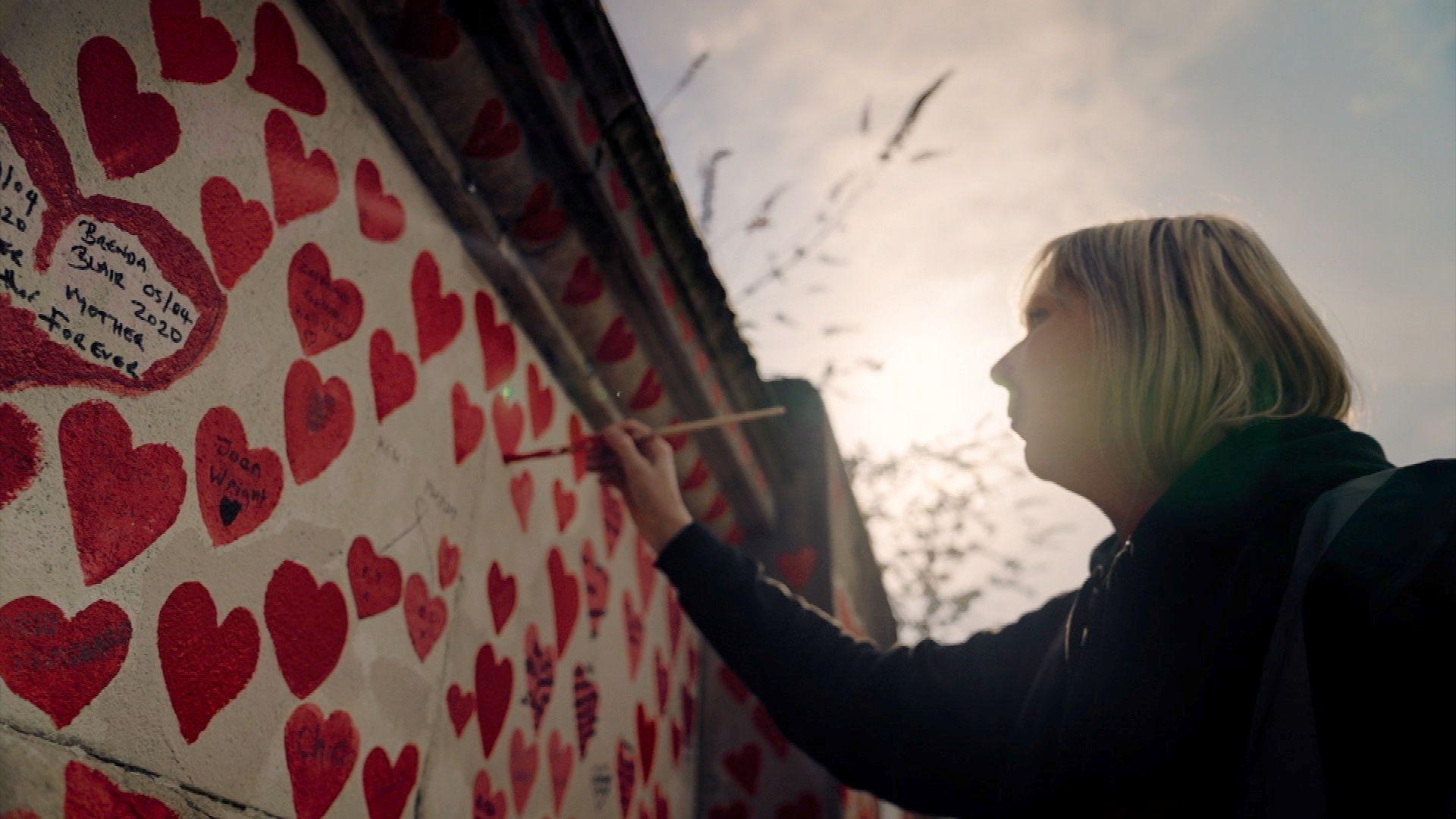 A woman, wearing a black coat and with short blonde hair, is painting a red heart on to the Covid memorial wall