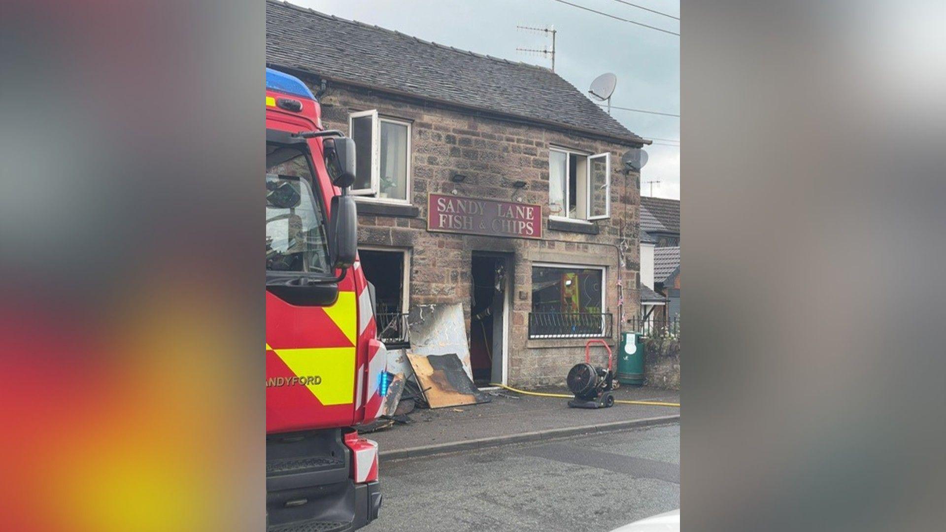 A brick building which has been badly burnt at the front. Charred wooden boards have been placed outside and a hose reel can be seen snaking inside through the open front door. A red sign above the front door says Sandy Lane Fish & Chips. The front of a fire engine is on the left.