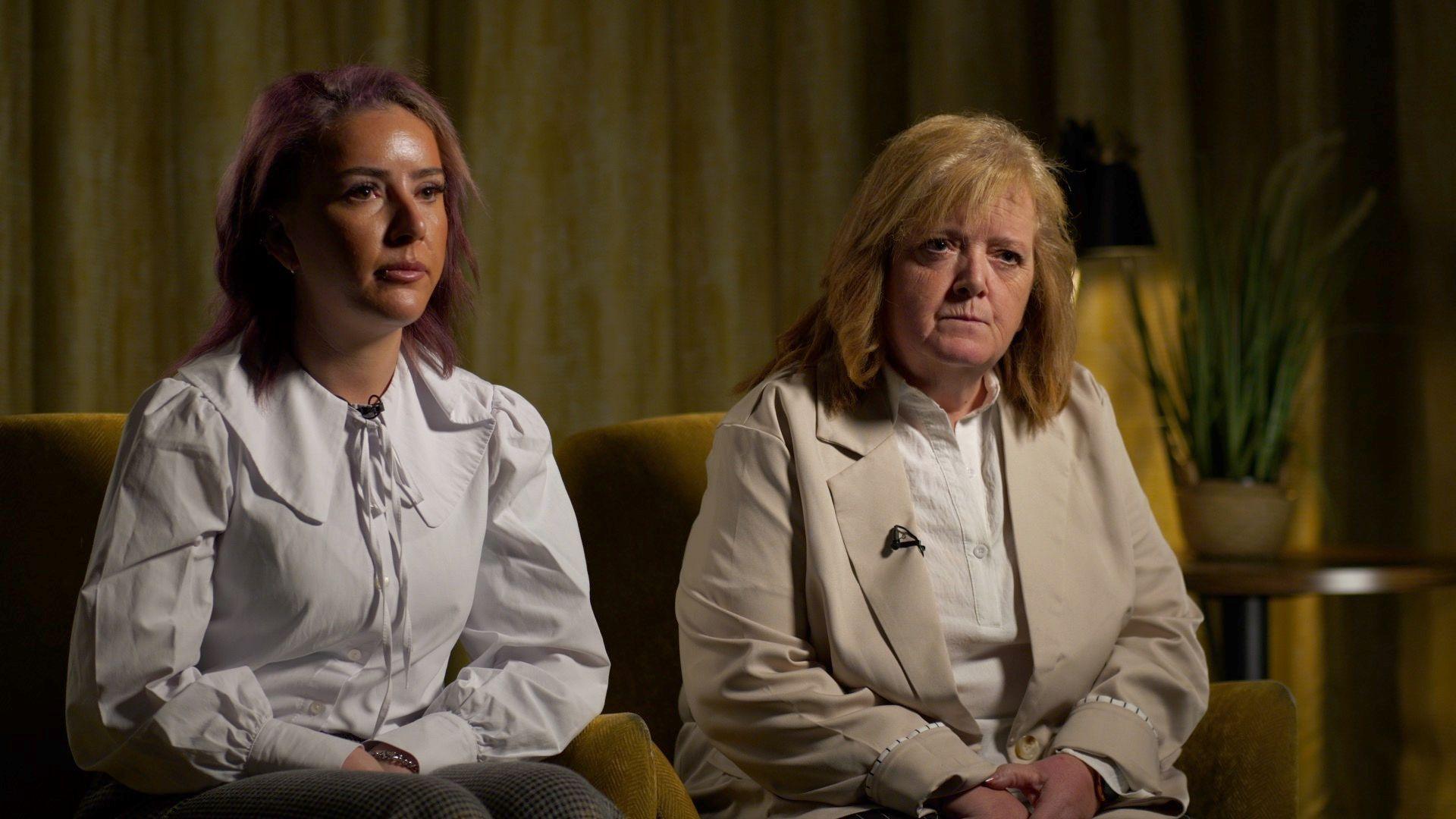 Two women are sitting on small yellow armchairs. On the left is a woman with purple-tinged hair, which is shoulder length, and she has tanned skin. She is wearing a white shirt with a large collar. Next to her is her mother, who has blonde shoulder-length hair, a cream-coloured smart coat and a white shirt. Both appear very solemn.