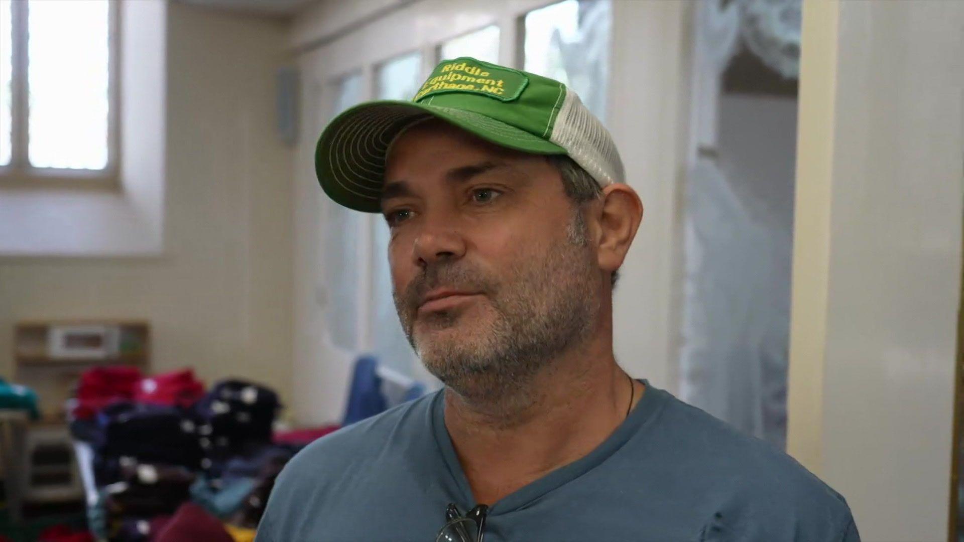 A man wearing a blue shirt and green and white cap. He is stood in a building with clothes on tables behind him.