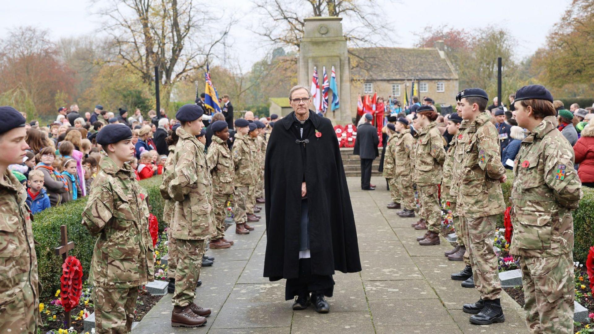 A member of the clergy, dressed in black, walks towards the camera flanked by military cadets wearing camouflage uniform and dark-blue berets. They are standing to attention in front of a war memorial. Regimental colours and poppy wreaths can be seen.