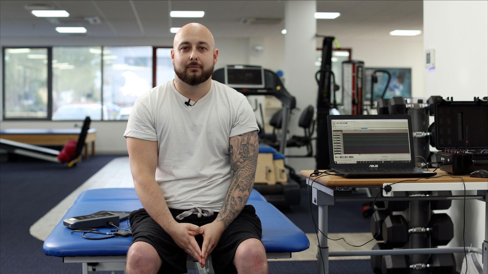 A bald man with tattoos on his left arms. He is sat on the end of a desk in an office with a computer next to him. He is looking at the camera while wearing a white t-shirt and black short