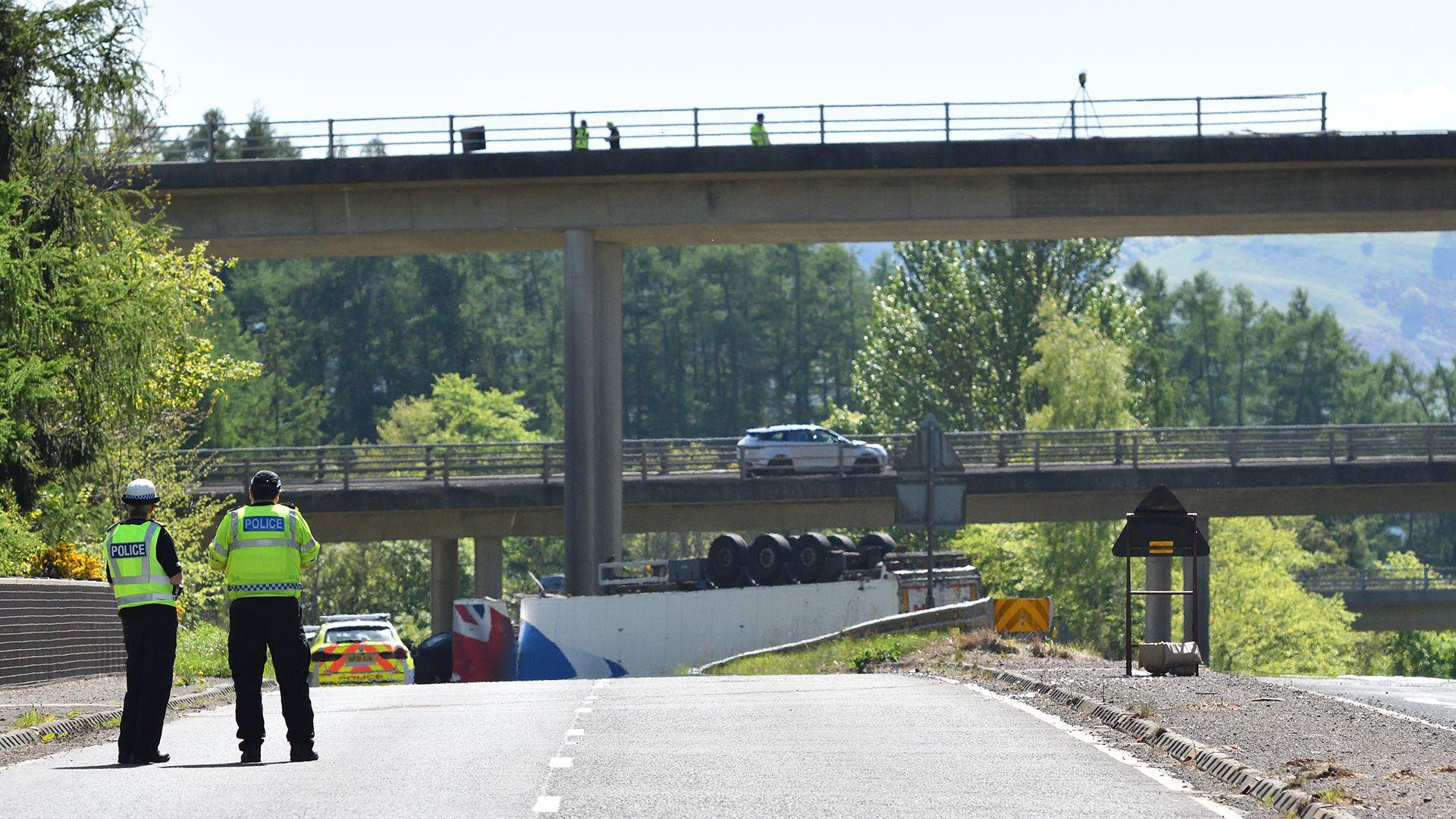 Two police officers stand in hi vis clothing bottom left of shot with an overturned lorry on the road in the distance below two concrete flyover bridges.