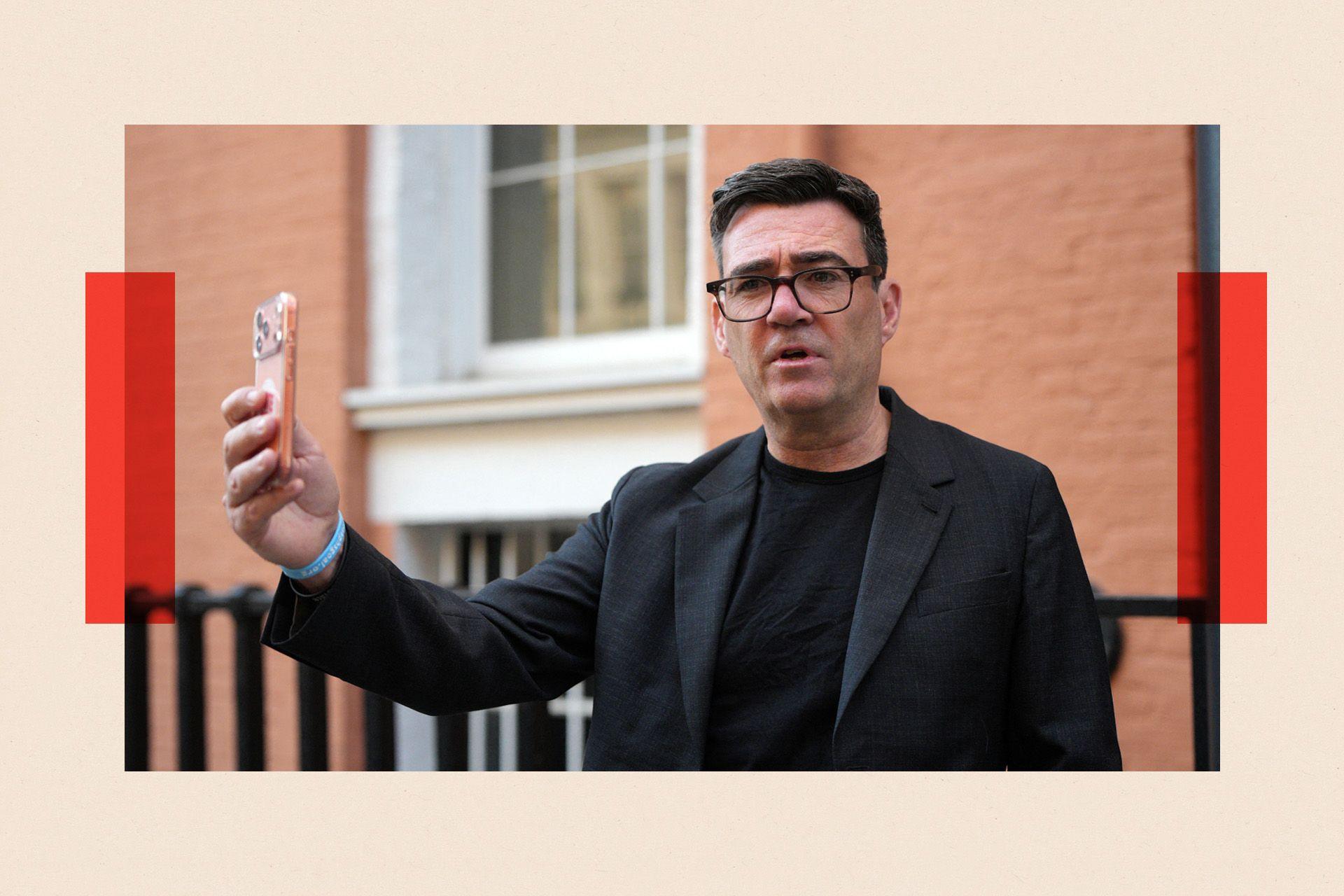Andy Burnham holds a peach-coloured smartphone as though taking a selfie. He has dark hair, dark-framed glasses and wears a dark jacket and T-shirt. Behind him is a red-brick wall.