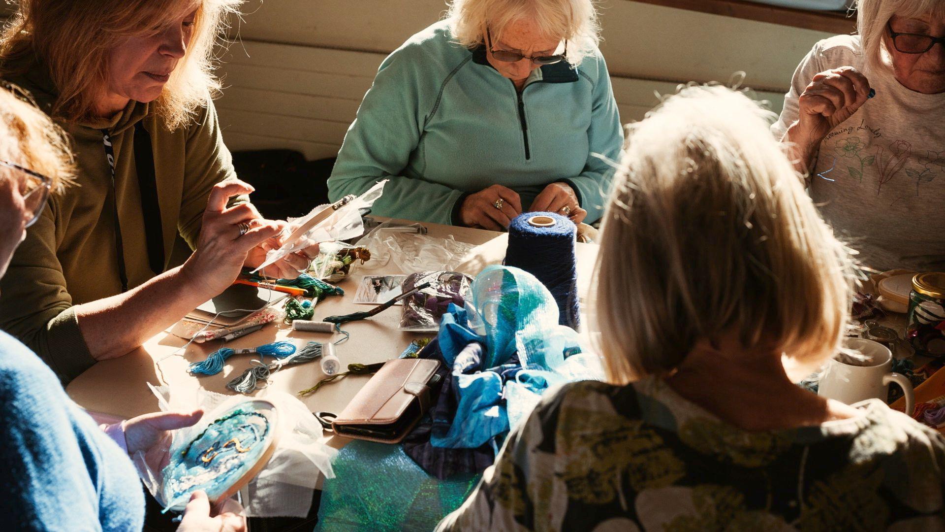 A group of five women are huddled around a table, working on their embroidery. There are various sewing materials on the table including threads, pieces of bright blue fabric, a phone and mugs of tea. The women are all looking down at their work, there is a yellow light coming in from the window giving the image a nice glow. 