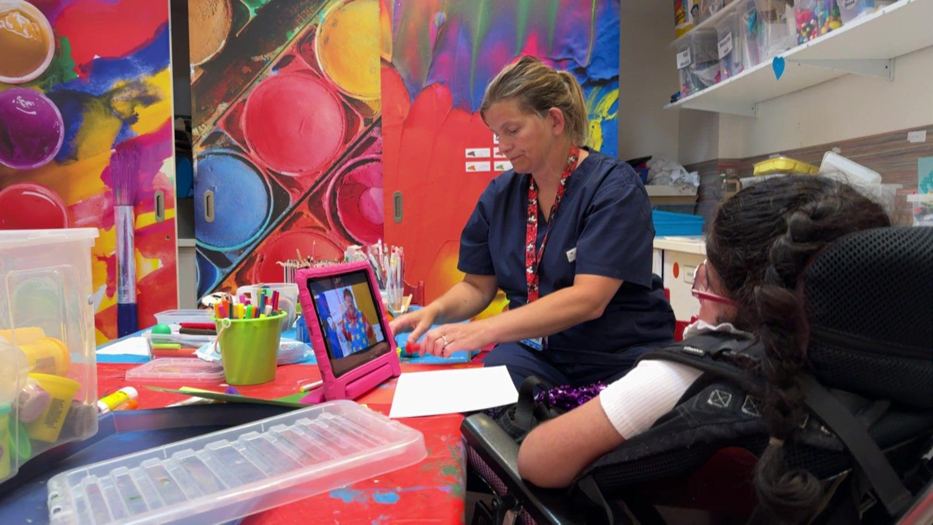 A woman siting at a desk in a colourful room full of toys and drawing materials next to a child that uses a wheelchair. The child is watching a programme on a tablet. The woman seems to be helping the child play with one of the toys.