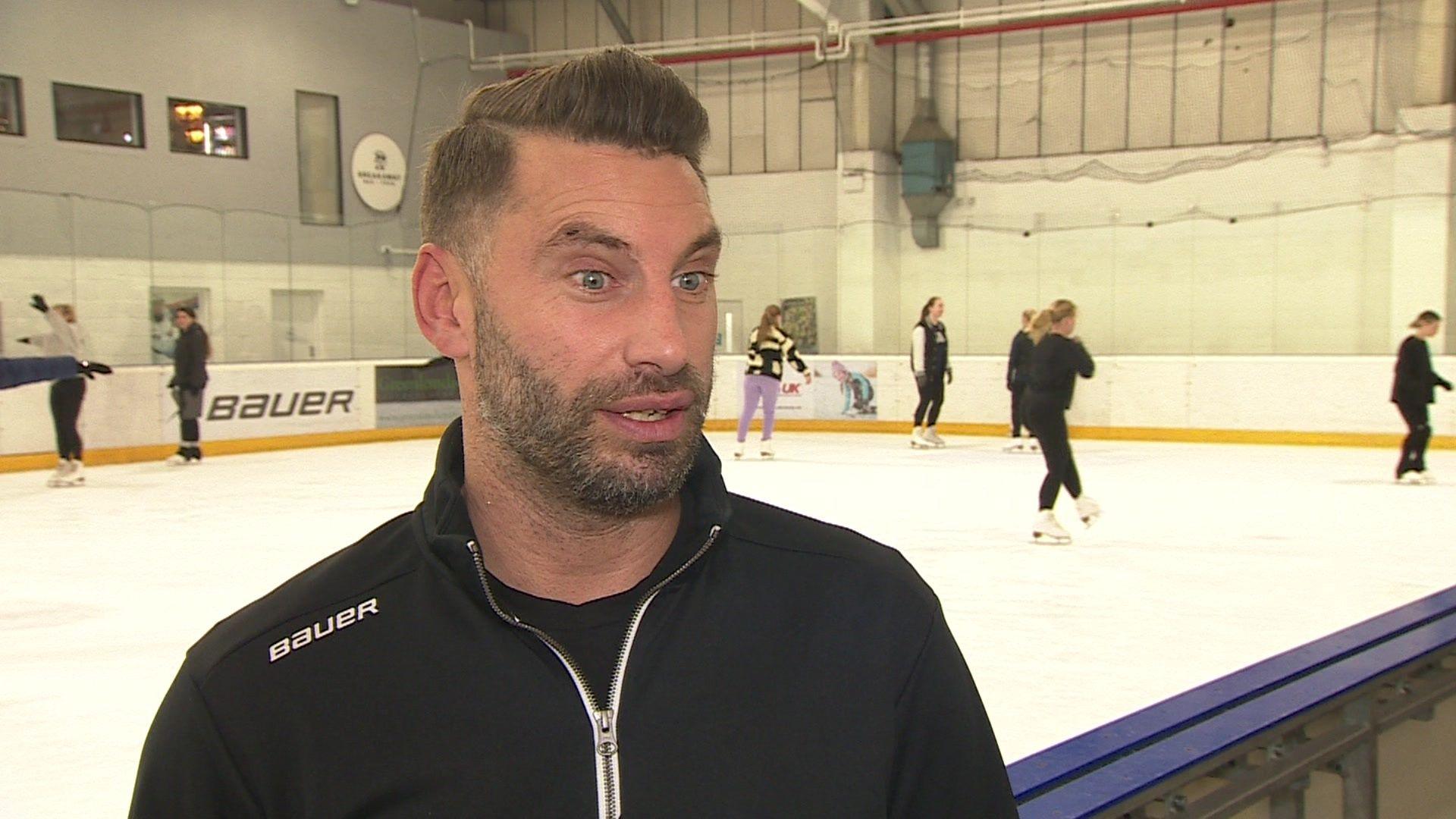 Matt Foord, from Ozone Ice Rink, he has dark hair and wears black. He has dark stubble. Behind him several people are skating on the indoor ice rink.