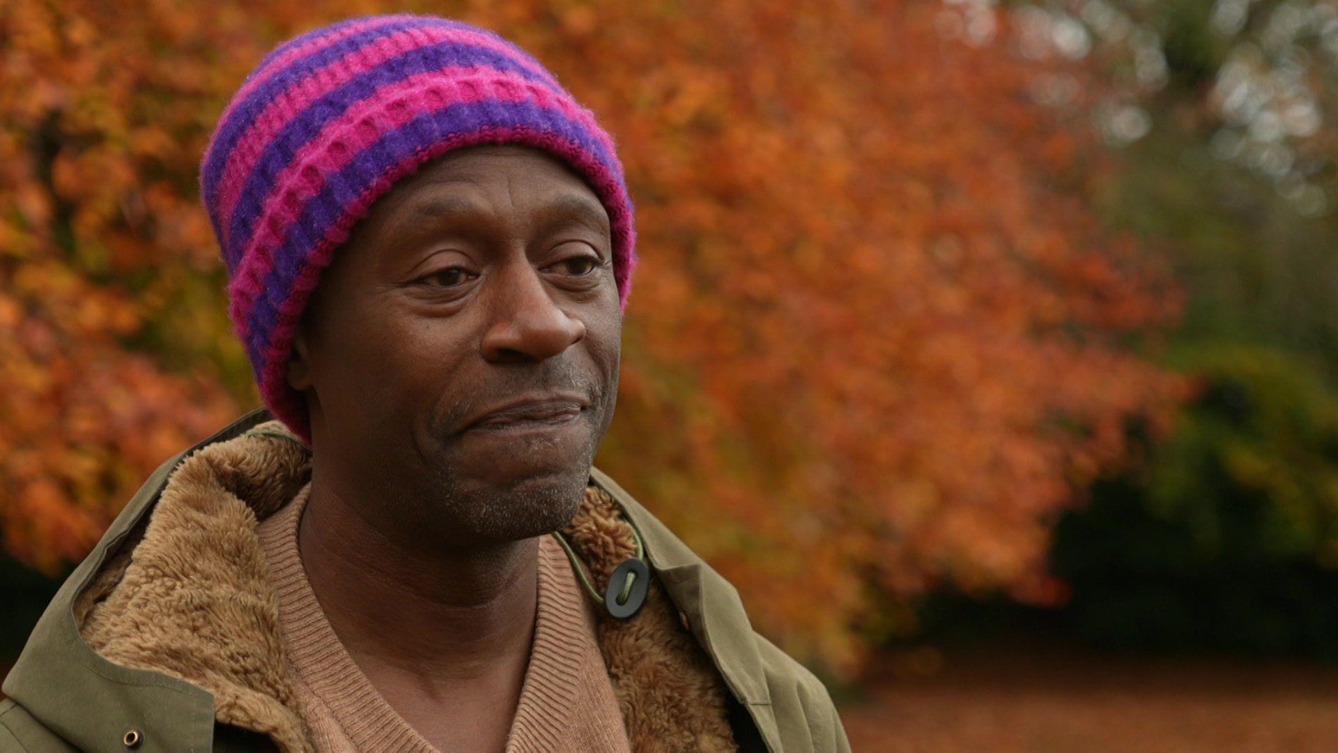 Mark Burnett stands in a park in autumn. He is wearing a purple and pink woolly hat and a green fur winter coat over a brown jumper.