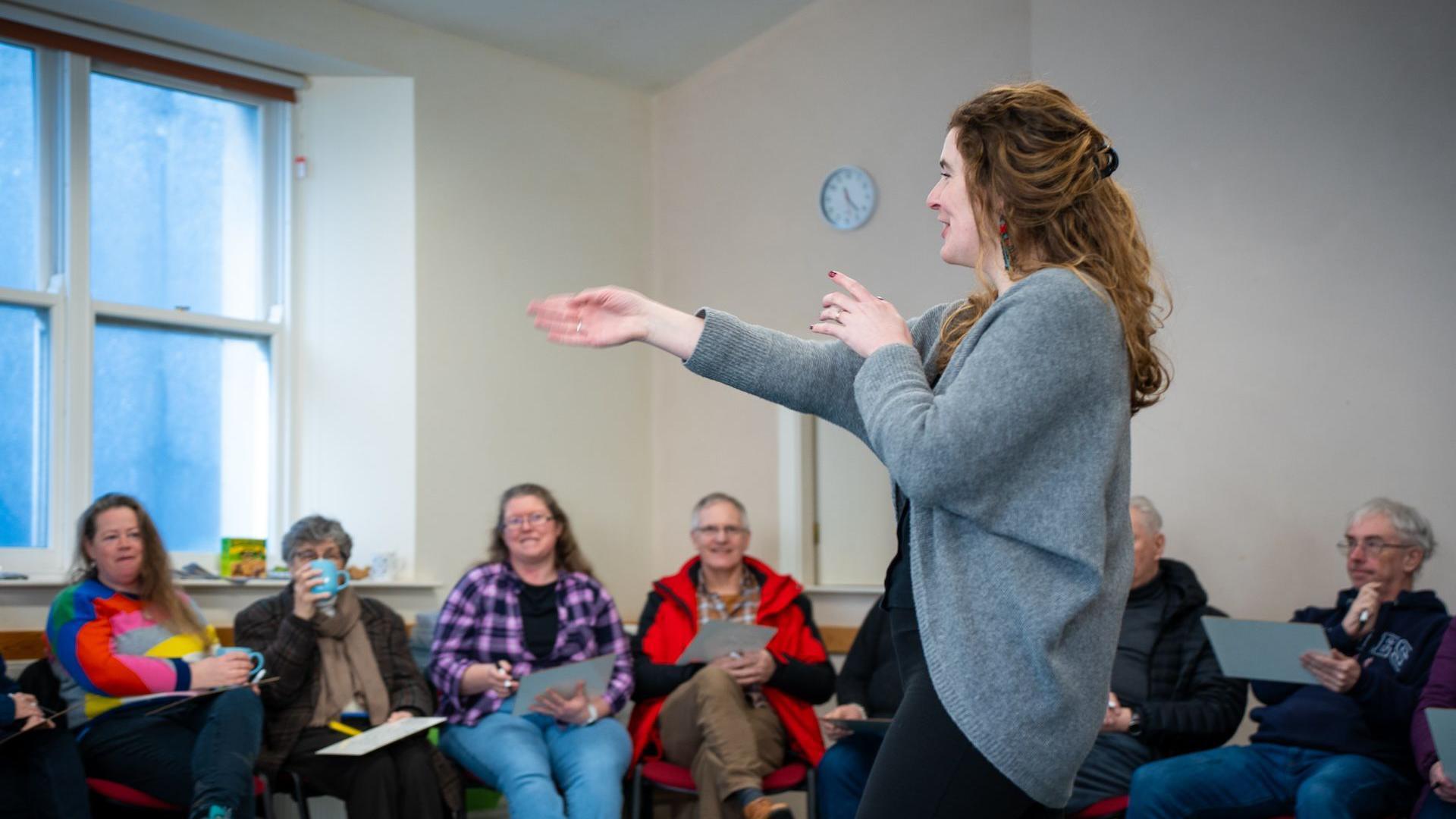 A woman with brown curly hair wearing a grey cargigan and pointing and standingl with 6 people watching her with notebooks.