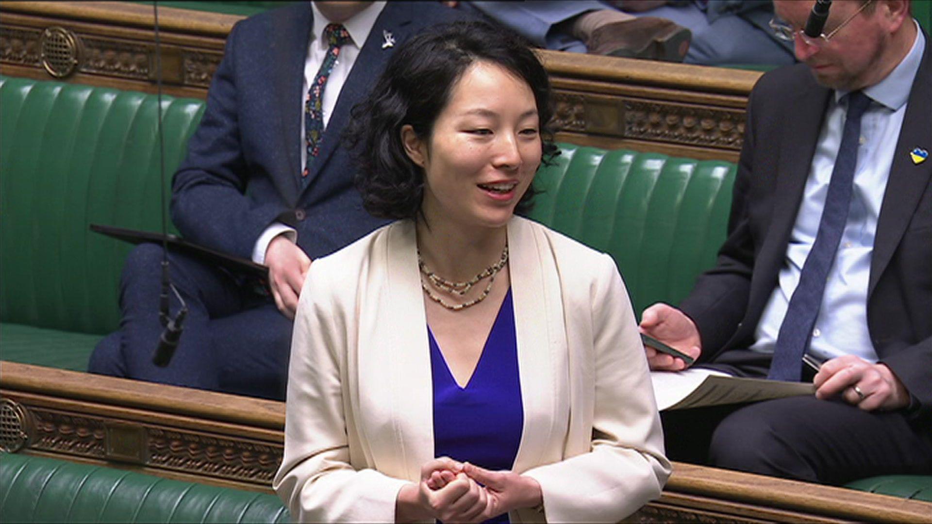 A woman with short black hair and wearing a blue top with a white blazer. She's standing up to speak in the House of Commons, with the green benches visible behind her.