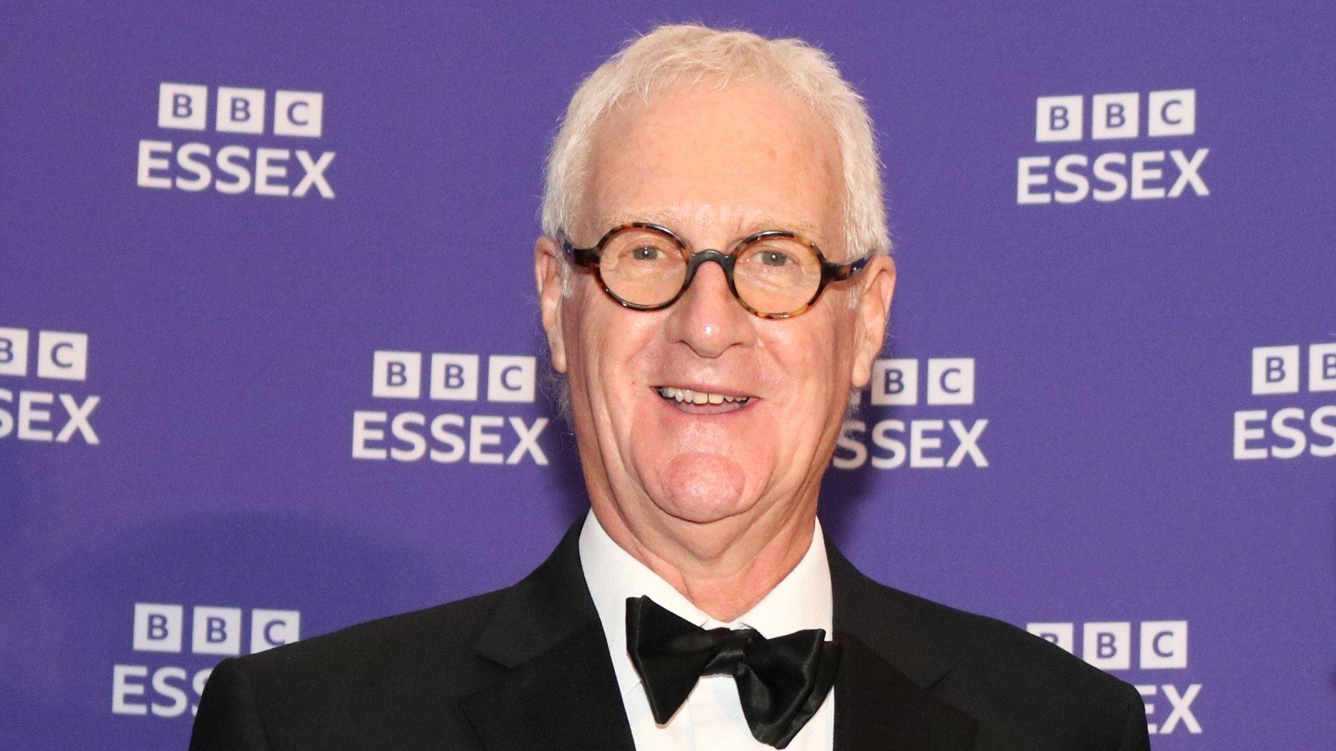 Dave Monk stands in front of a purple backdrop with several white BBC Essex logos. Dave is wearing a tuxedo jacket, white shirt and black bow tie. He has grey hair, is smiling at the camera and wears round spectacles.
