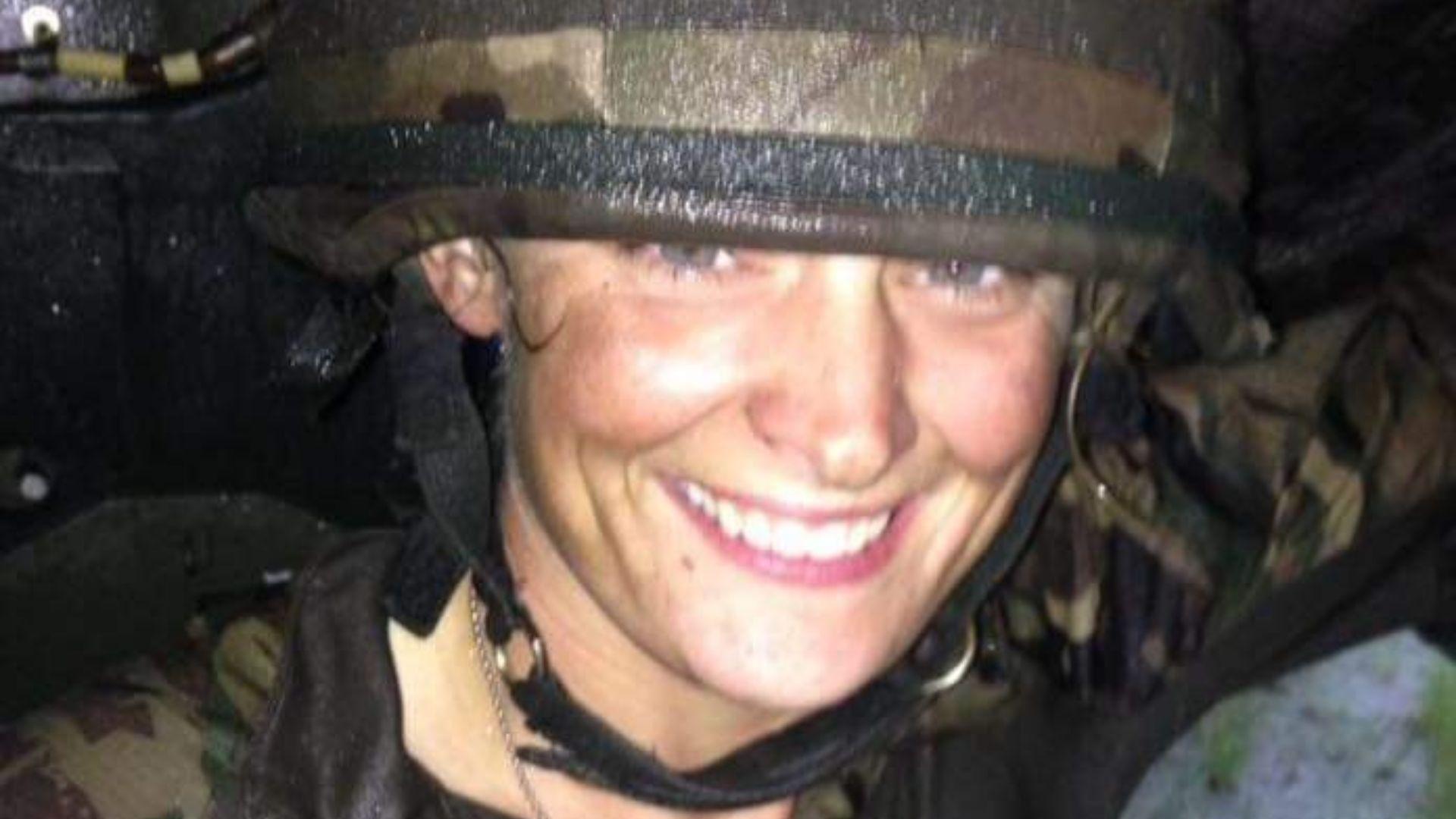 A head and shoulders shot of a female soldier in a military helmet and camouflage long-sleeve top. She is smiling at the camera. Her face appears slightly dirty, as if she has been training, and her clothing appears wet. The bright flash is illuminating her face but the photograph appears to have been taken at night. 