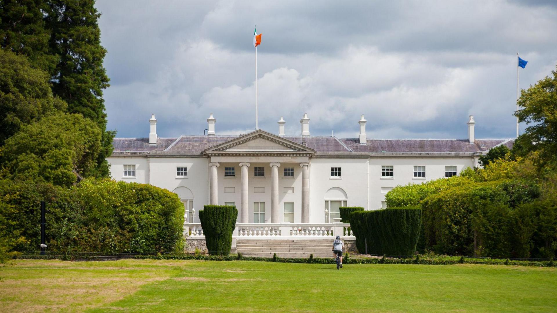 An exterior shot of Áras an Uachtaráin. It shows a two-storey white building, resembling the White House in the USA. There is a flag pole with the Irish tricolour on it at full mast. Surrounding the building are large trees, bushes and a large patch of grass.