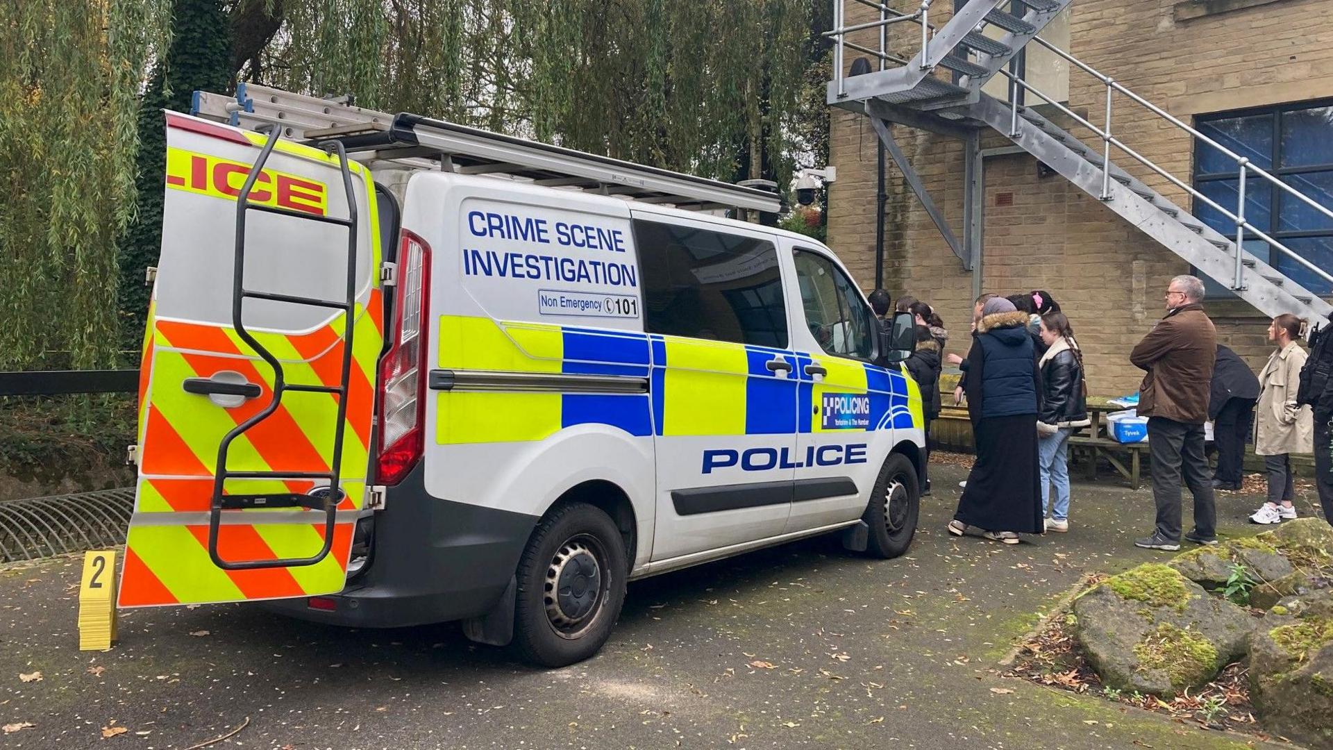 The back of a police Crime Scene Investigation van with one of its rear doors open and the word "police" in blue lettering down the side of a white panel. Pupils and a teacher stand in the background. 