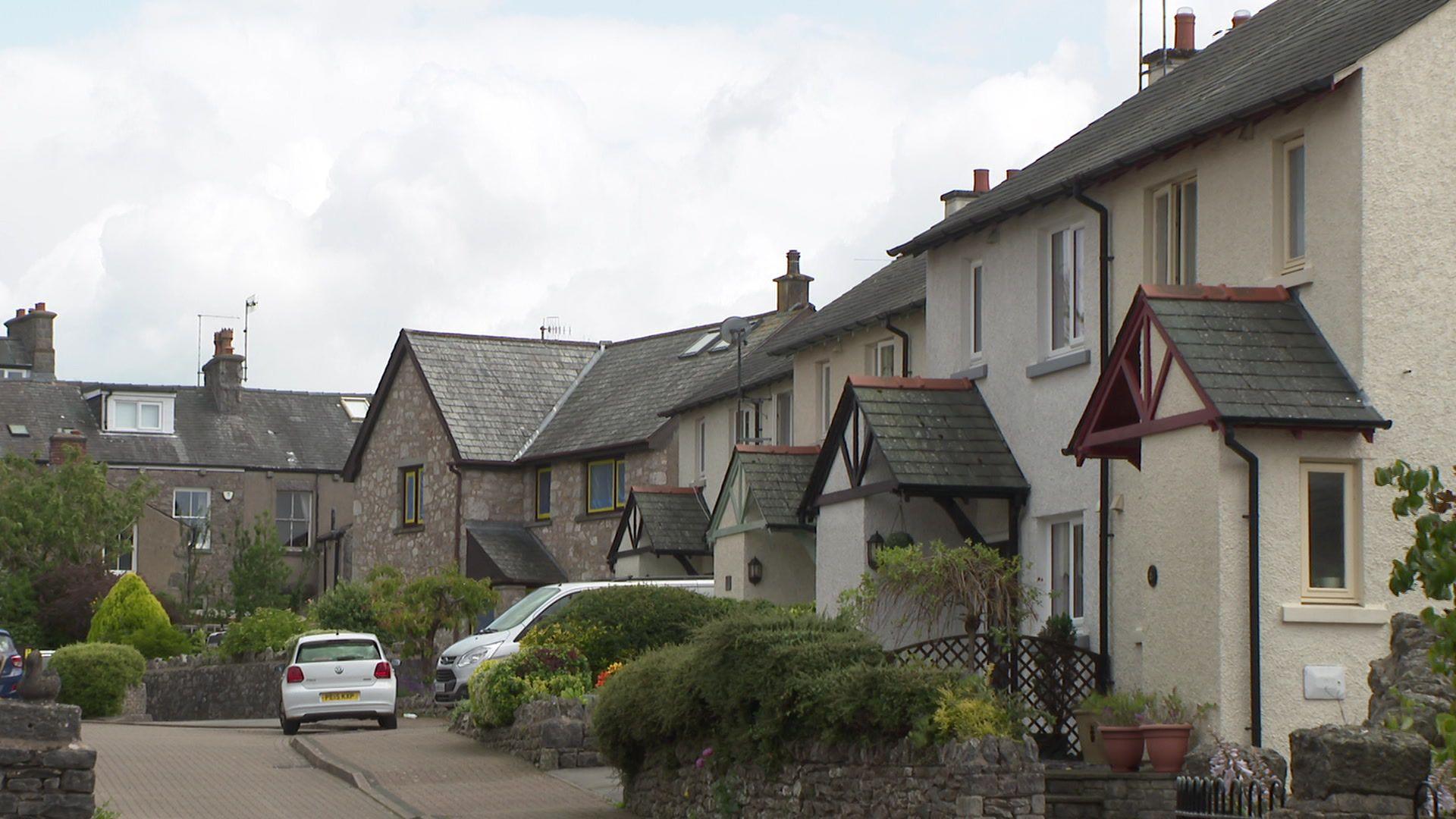 Semi-detached and detached houses line this quiet street in Silverdale. Potted plants and bushes are visible in the front gardens of many of the homes.